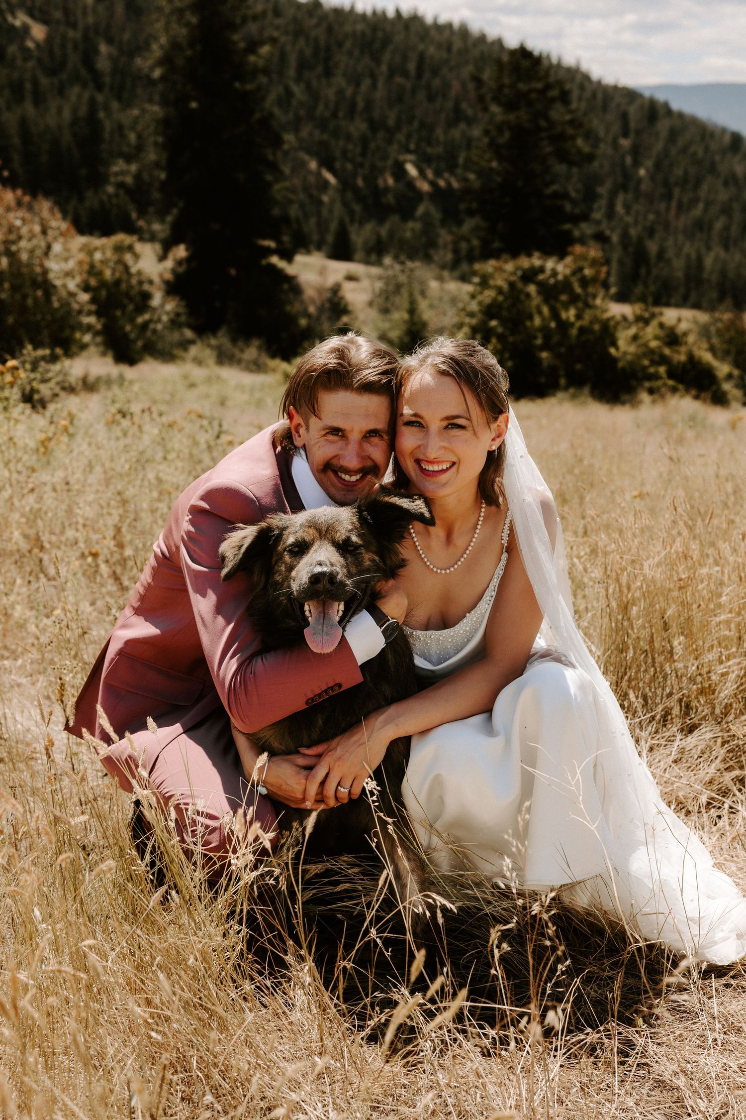 A couple and their dog share joyful moments together in a sunny meadow with mountains in the background.