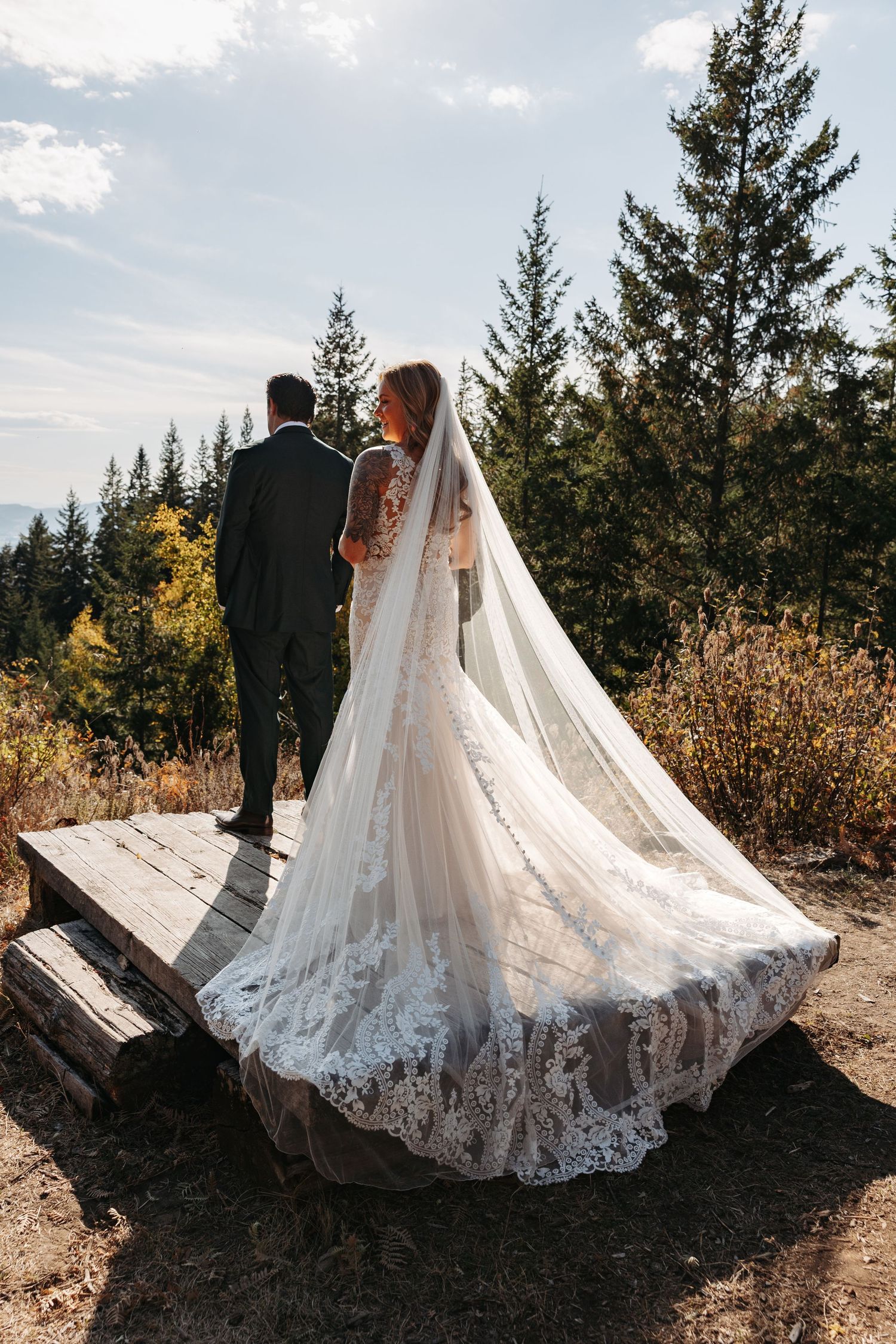 A wedding couple stands on a wooden platform surrounded by evergreen trees as the bride's lace train flows behind her.