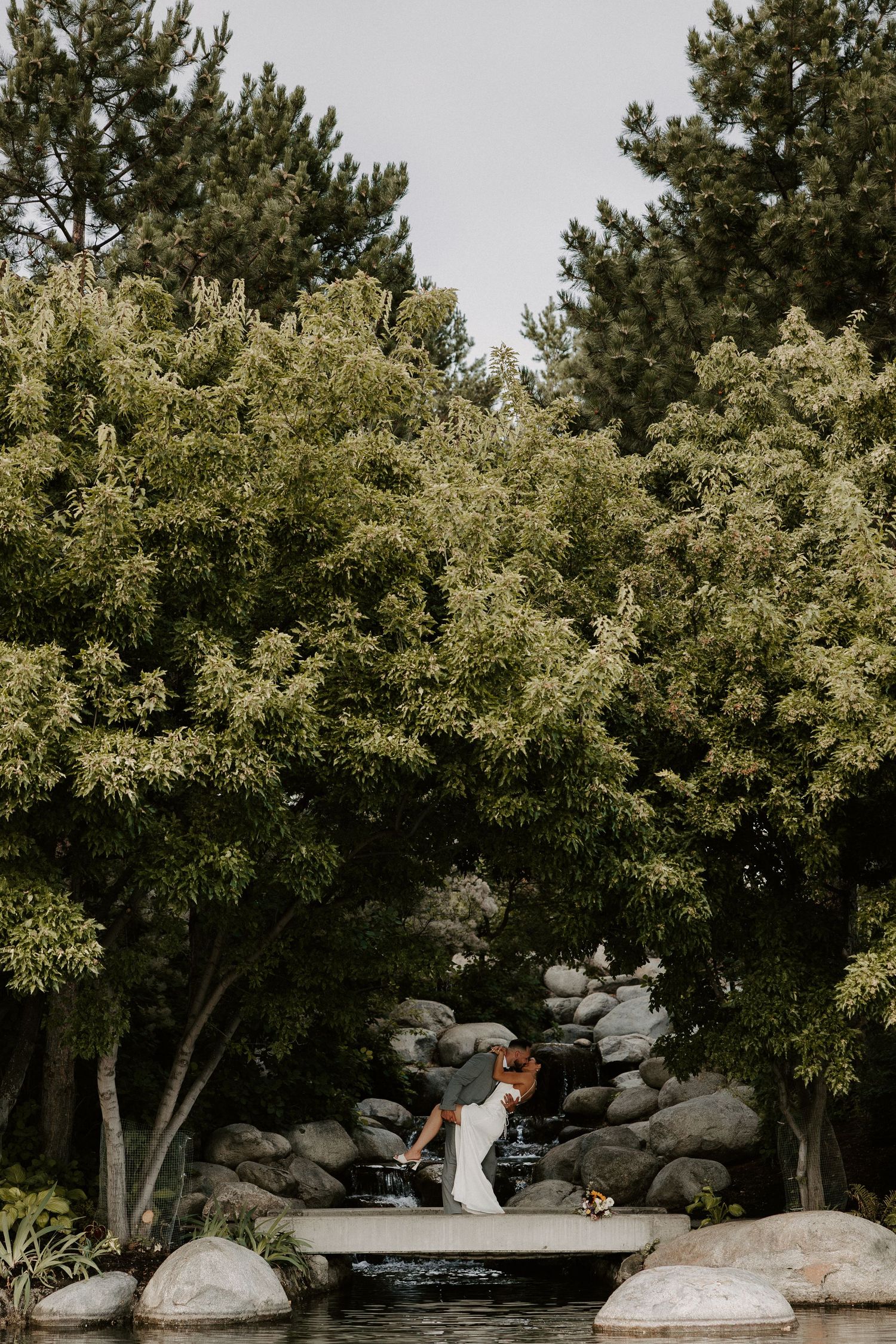 Couple standing on small wooden bridge over rocky creek surrounded by lush green trees in romantic outdoor setting.