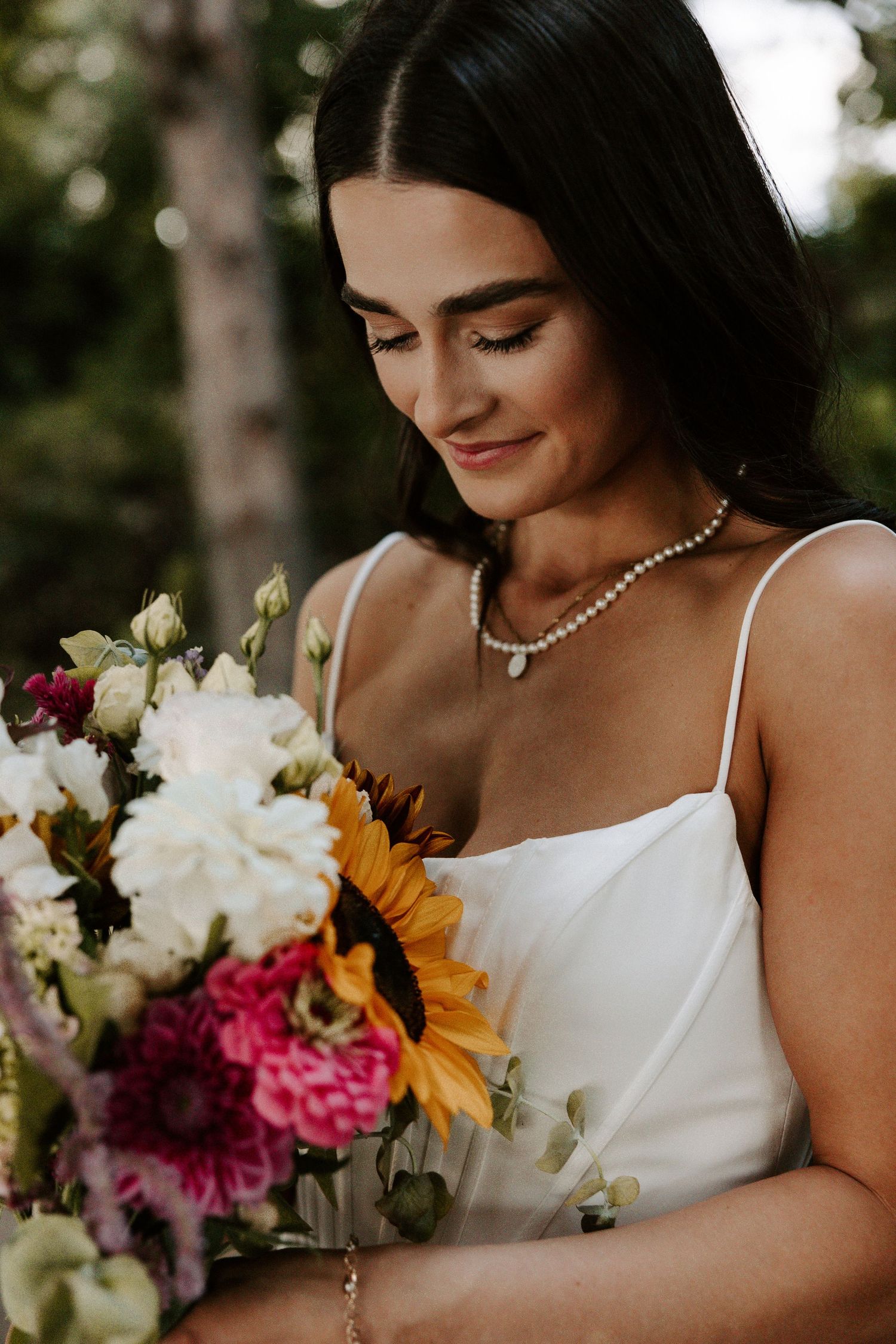 A bride in a white gown admires her colorful wedding bouquet featuring sunflowers, pink blooms and white florals.