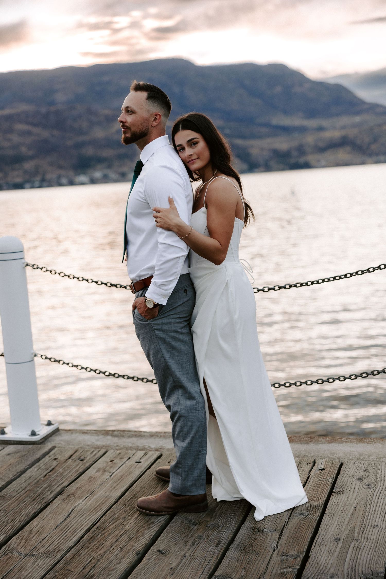 A couple in wedding attire embraces on a dock by the lake with mountains in the background at sunset.