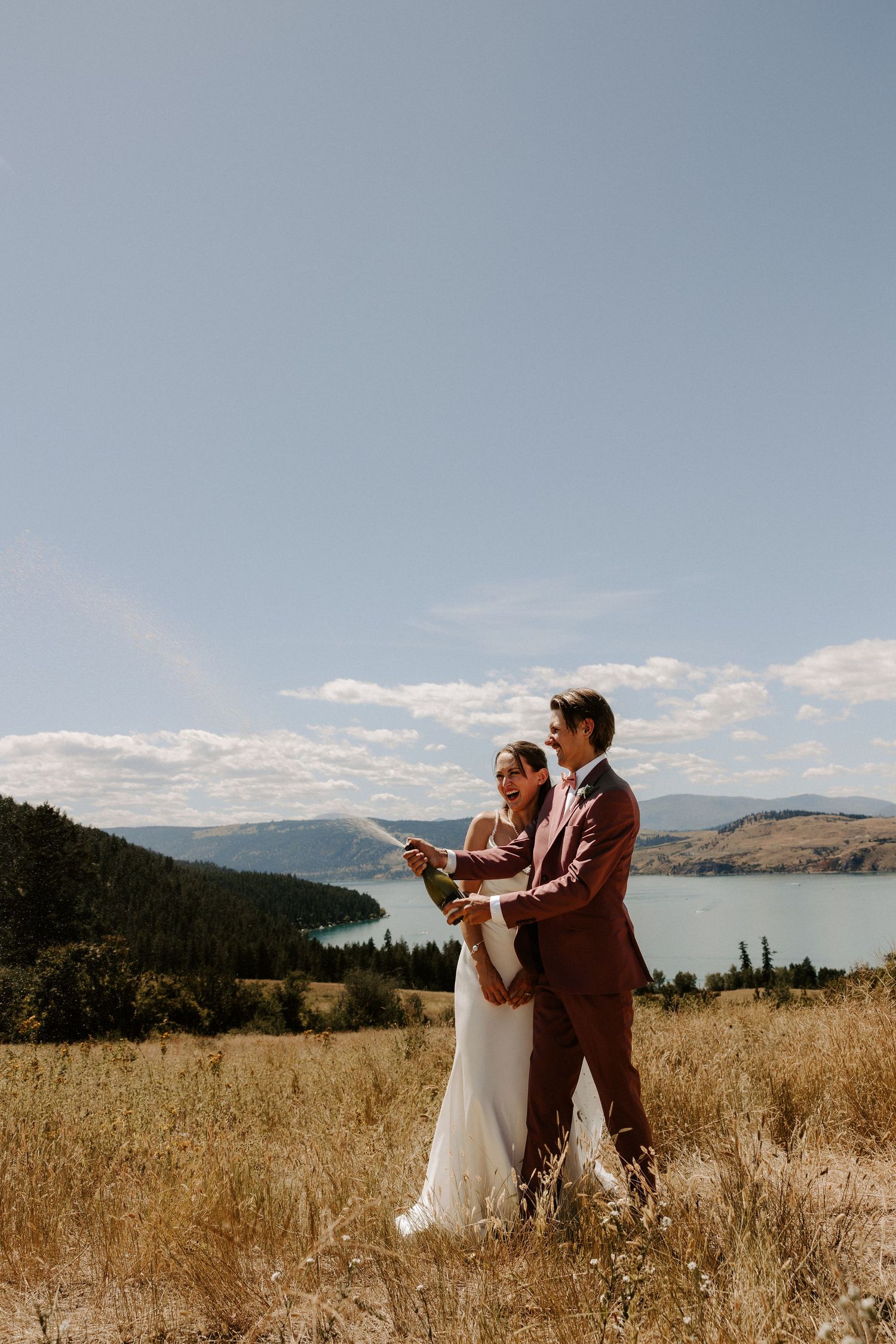 A couple in wedding attire stands together on a hilltop overlooking a scenic lake and mountains on a sunny day.