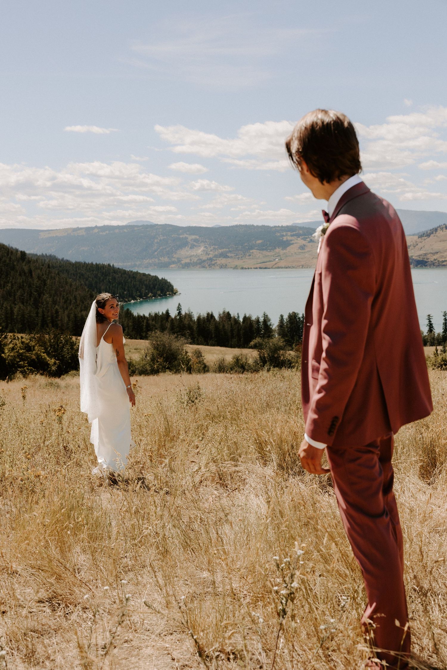 A couple in wedding attire stands in a golden field overlooking a scenic mountain lake view.