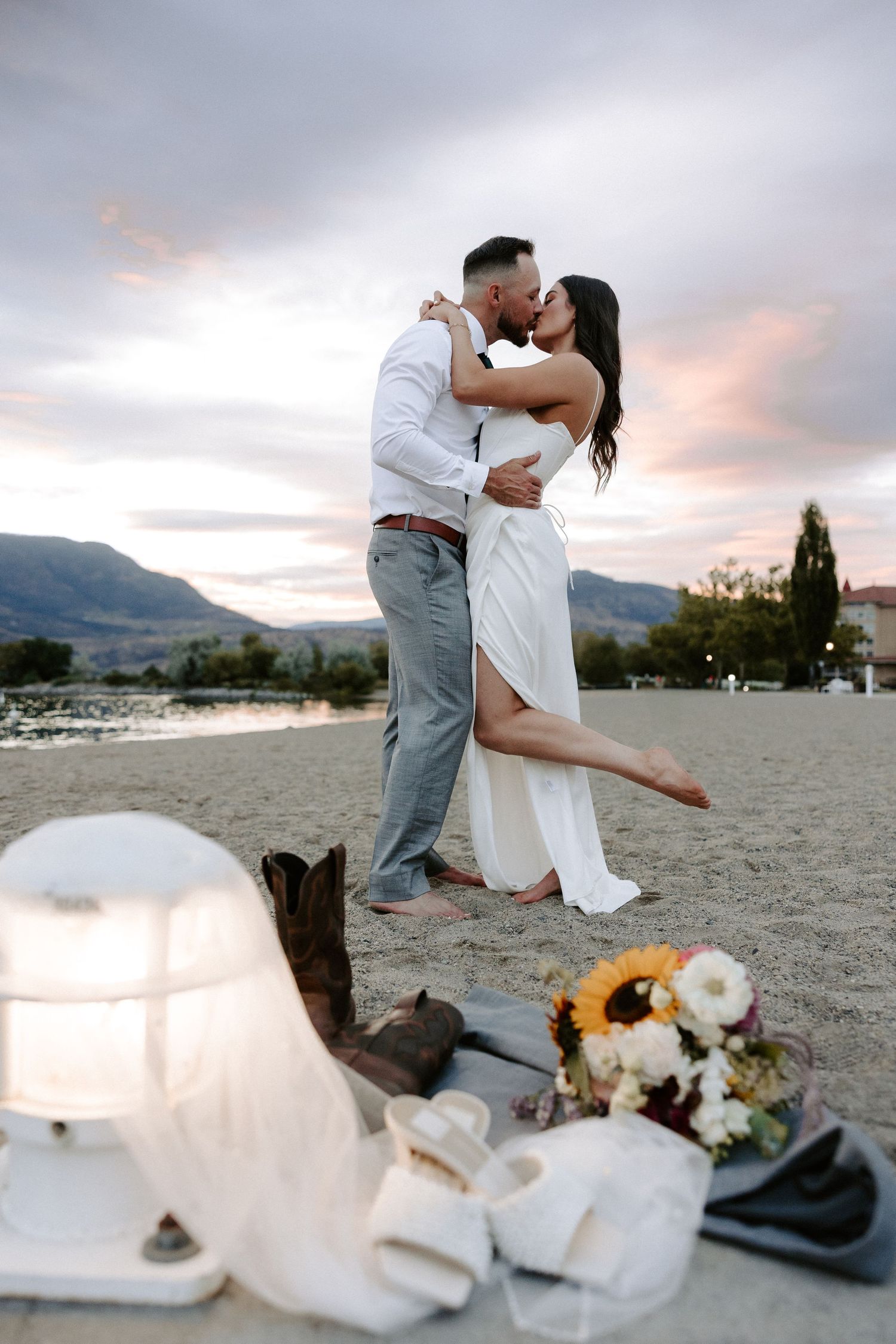 Romantic beach moment at sunset with cowboy boots and sunflowers laying in the sand as couple embraces.