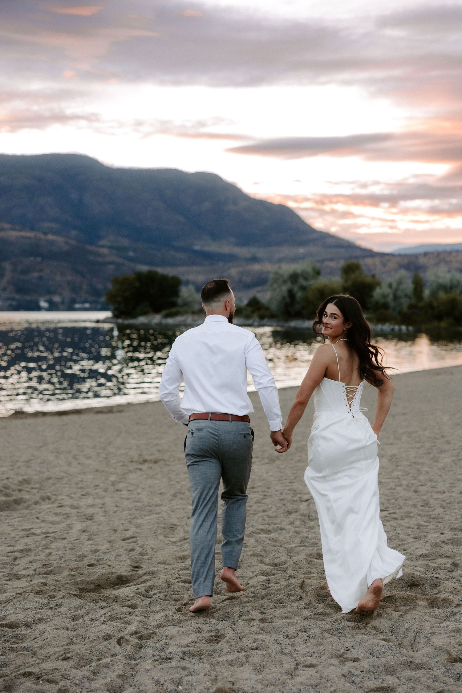 A couple walks hand in hand along a beach with mountains and sunset in the background.