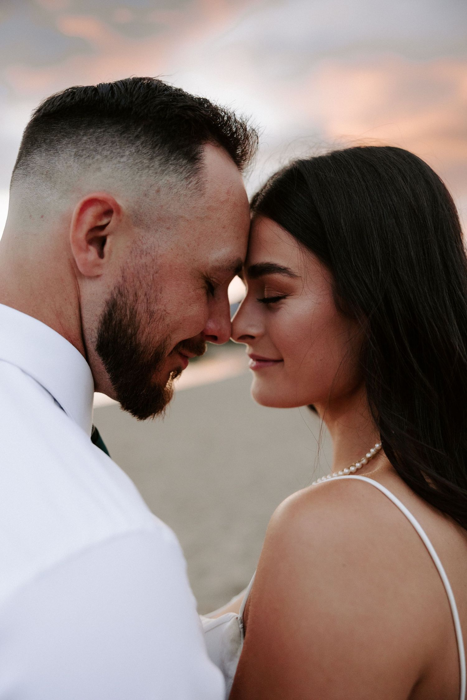 Intimate close-up of a romantic couple touching foreheads during sunset with cloudy pink skies in the background.