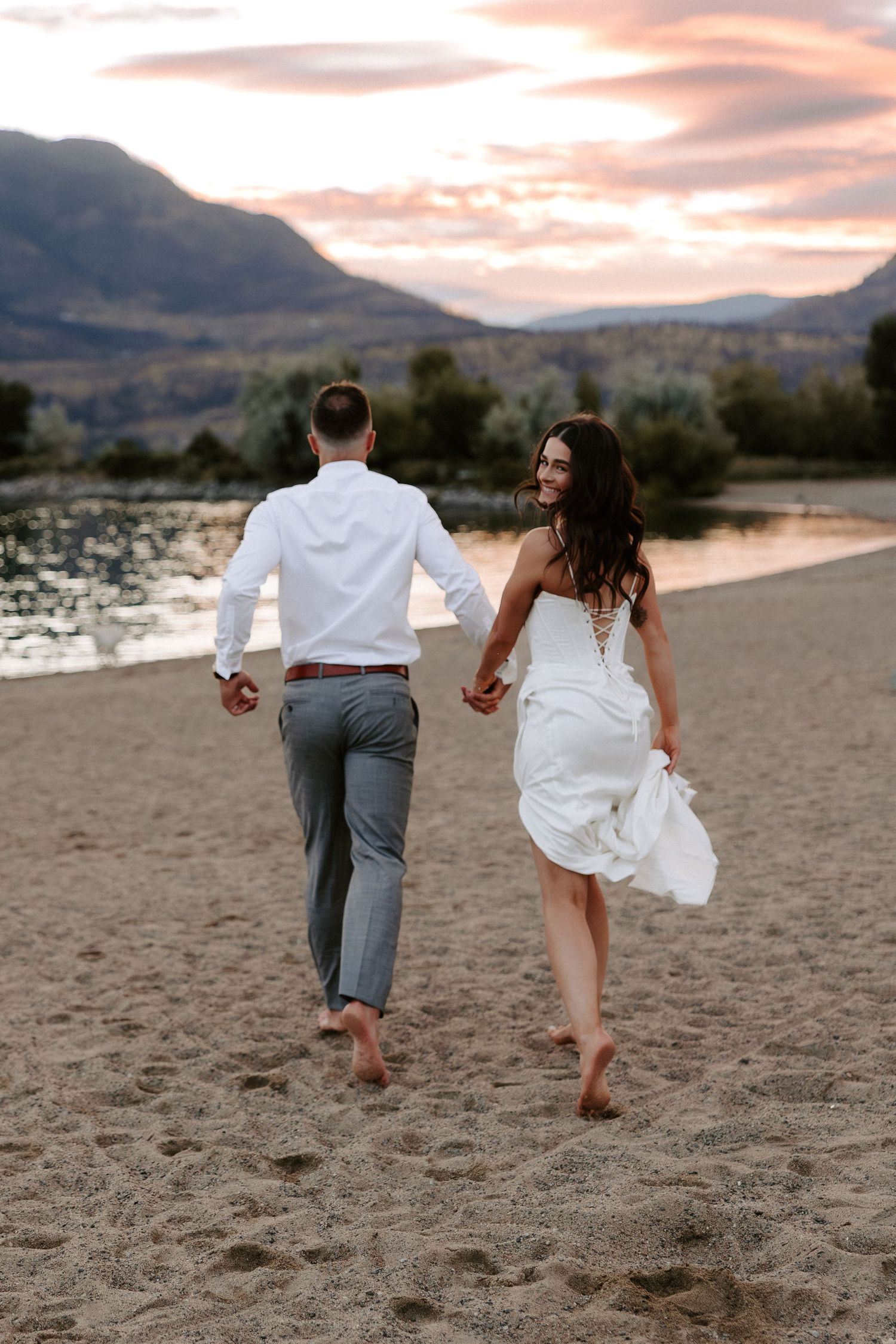 A couple runs playfully along a beach at sunset with mountains in the background, holding hands while kicking up sand.