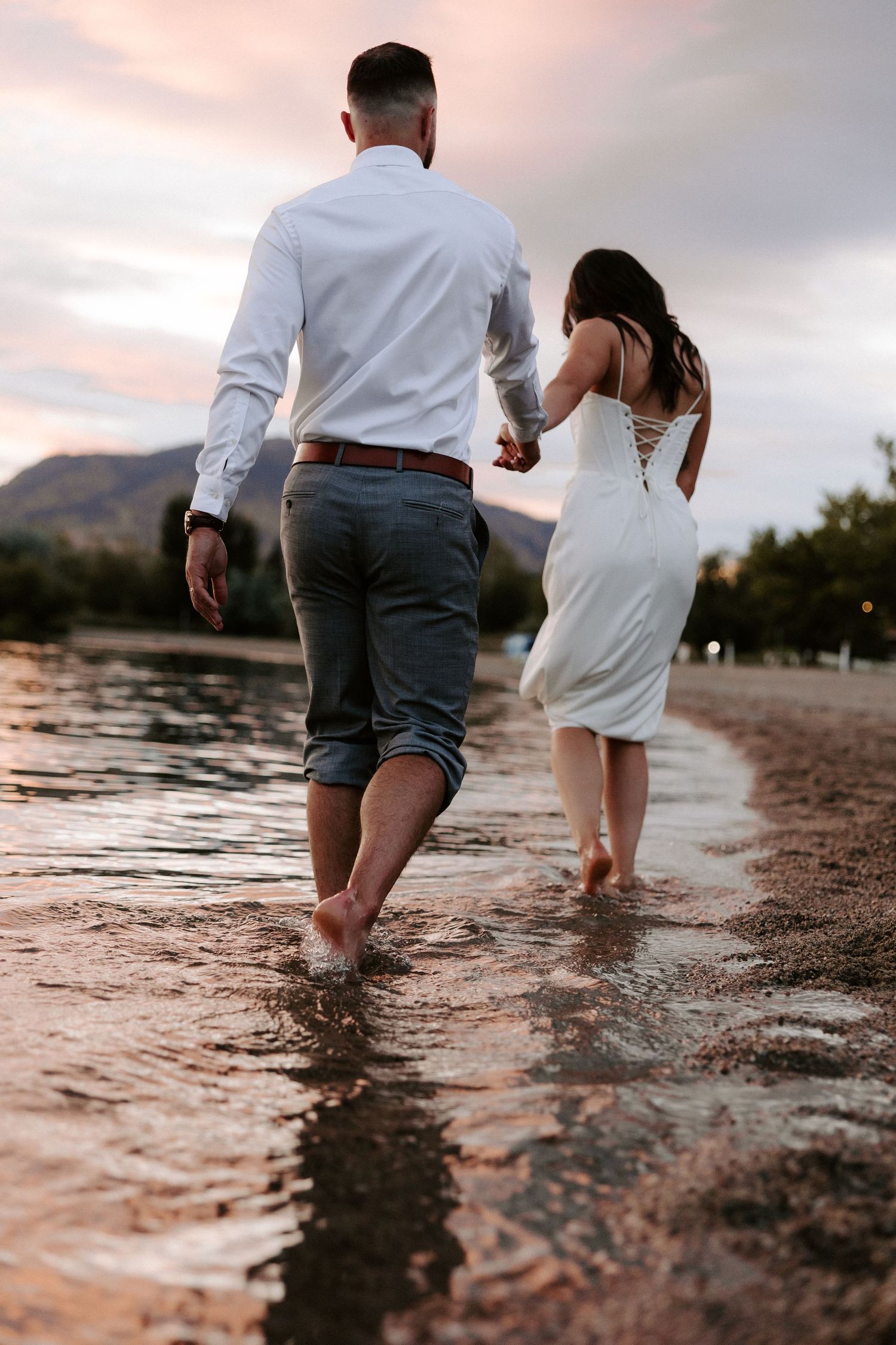 Couple walking hand in hand along the water's edge at sunset with mountains in distance.