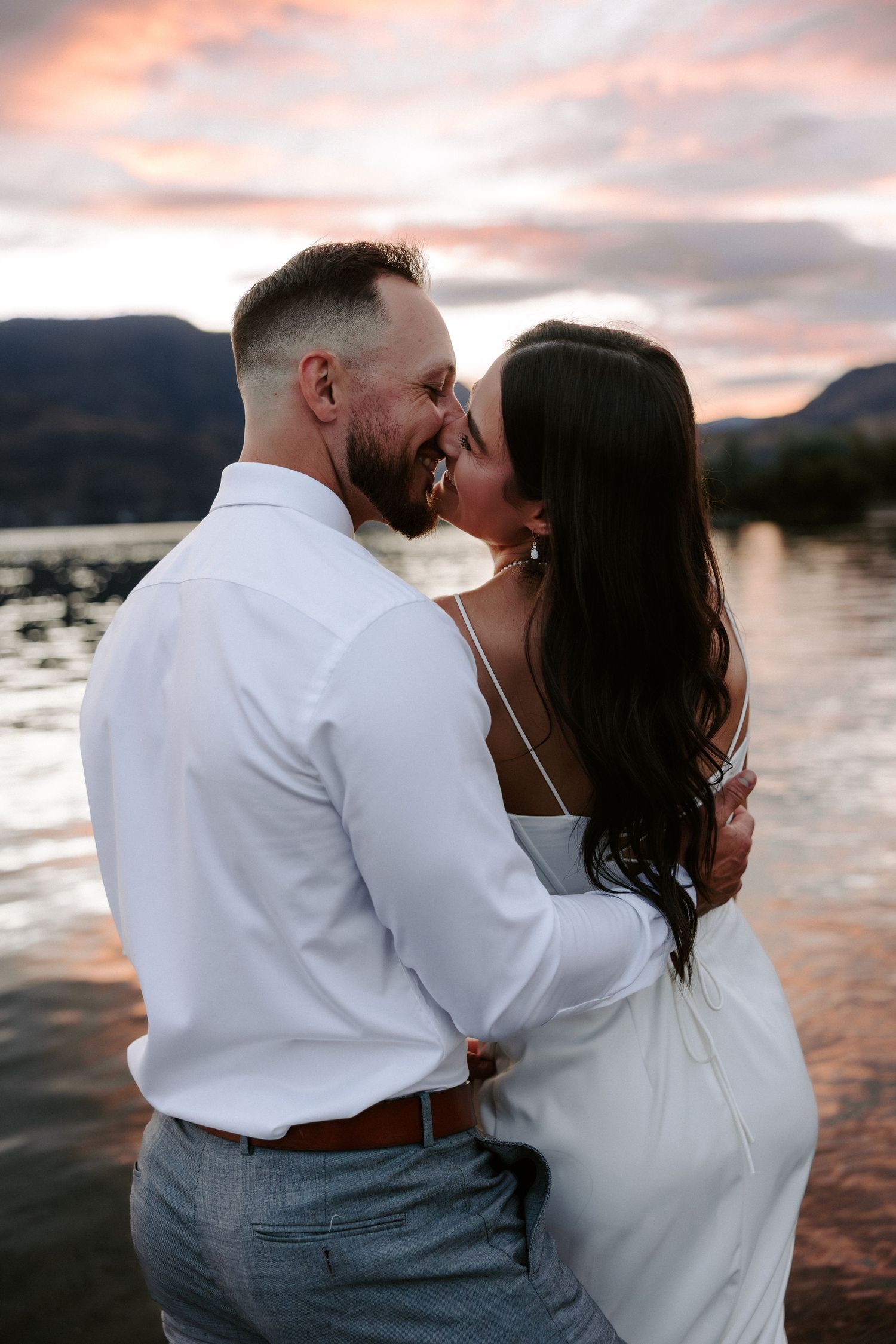 A romantic couple embraces while watching the sunset over a tranquil lake, creating a serene silhouette against pink sky.
