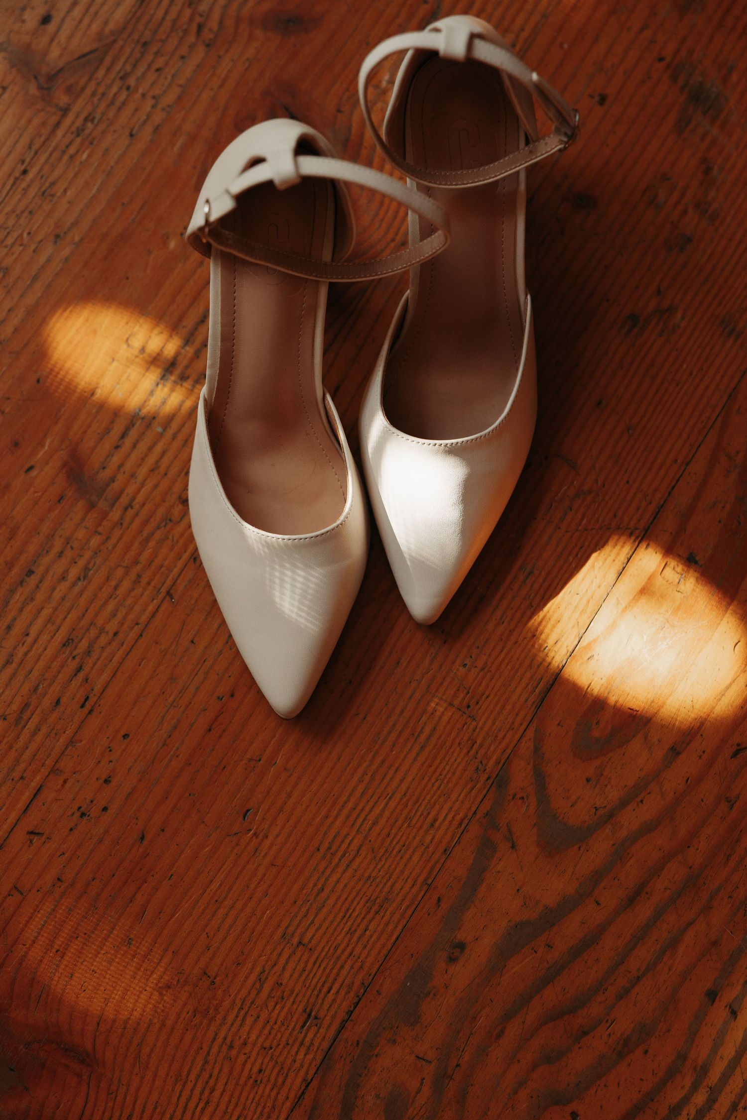 A pair of elegant white leather ankle-strap heels sits on a rustic wooden floor in warm lighting.