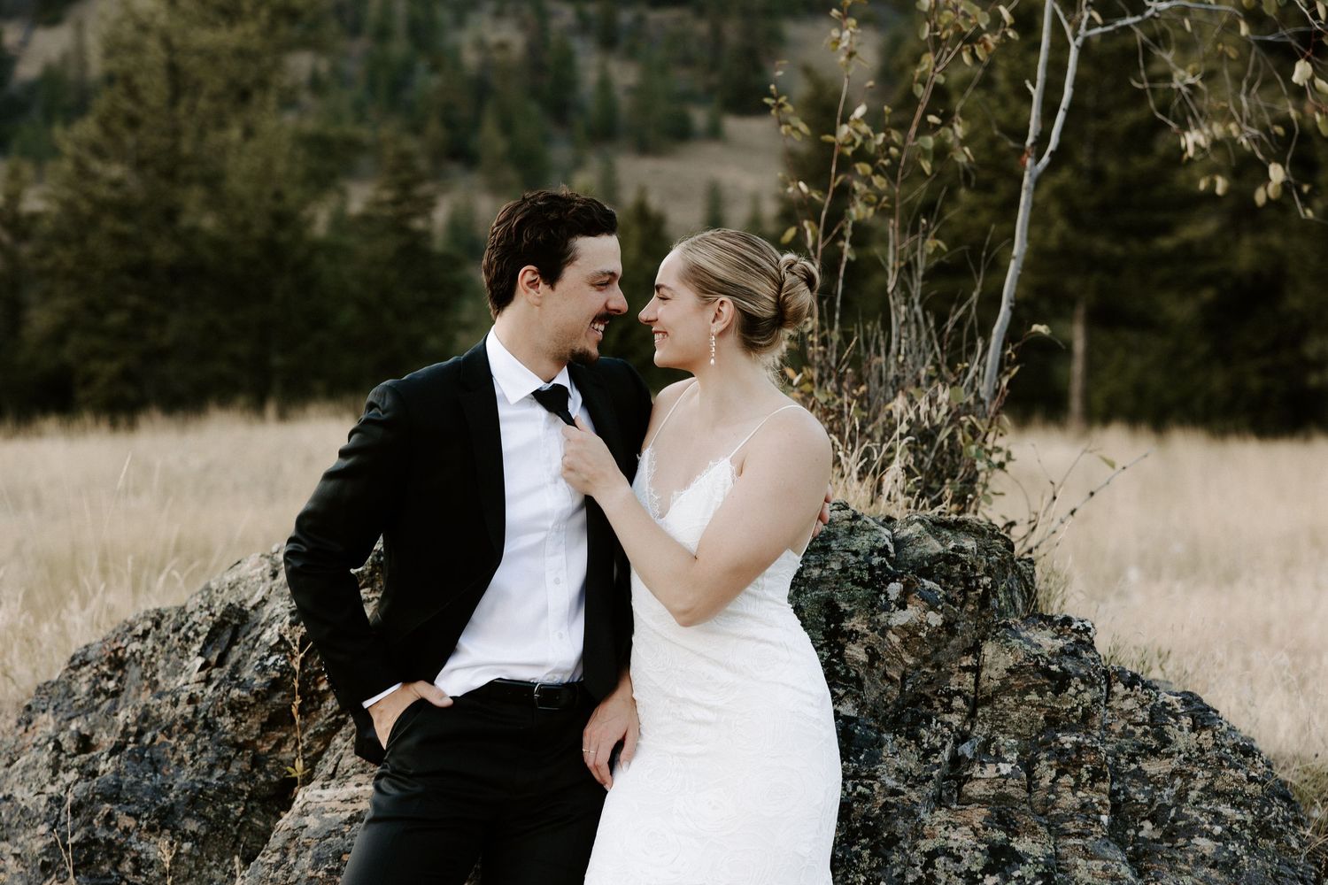 A romantic newlywed couple poses outdoors near rugged black rocks in a field at sundown.