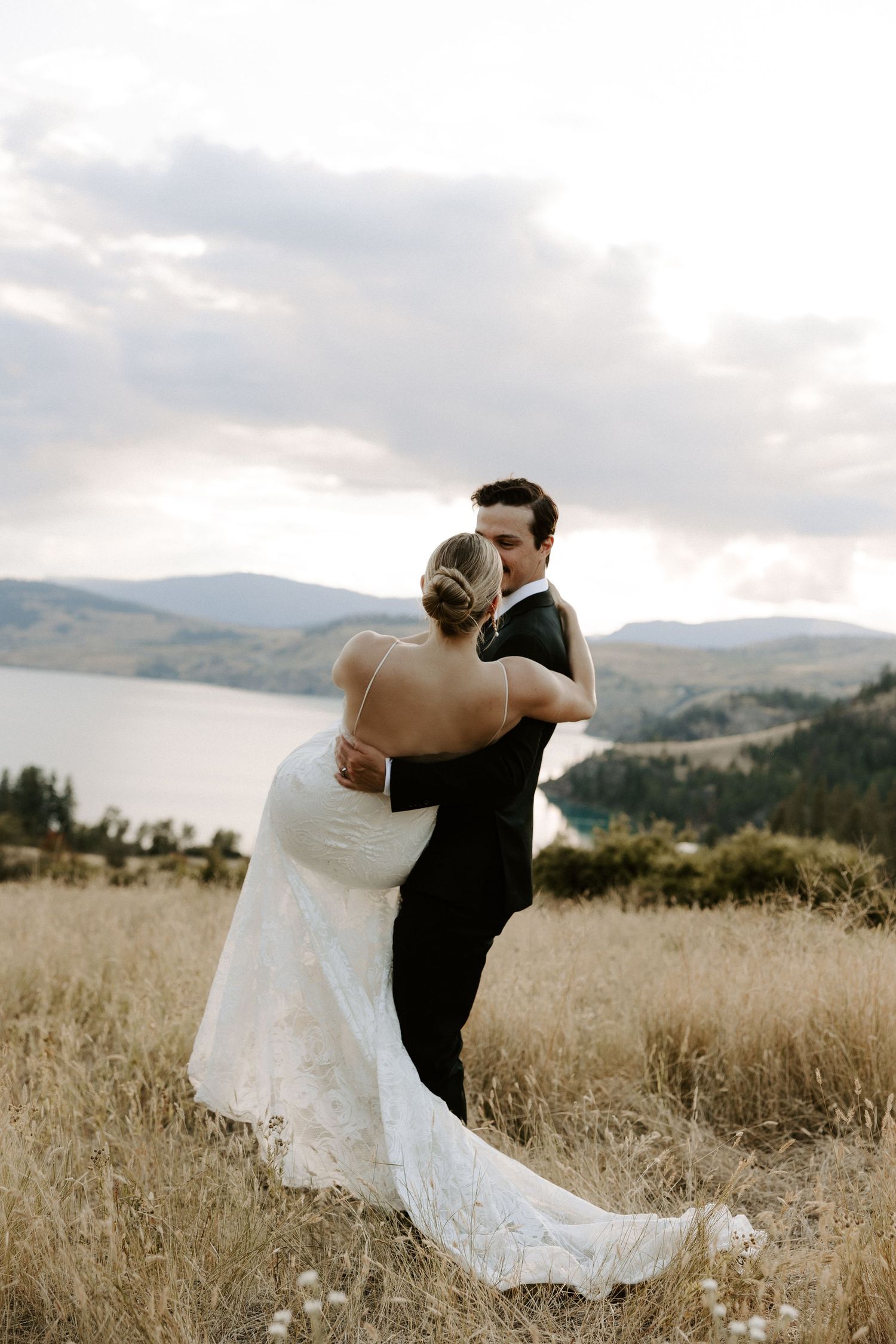 A couple shares their first dance as newlyweds on a hilltop overlooking a scenic mountain lake at sunset.