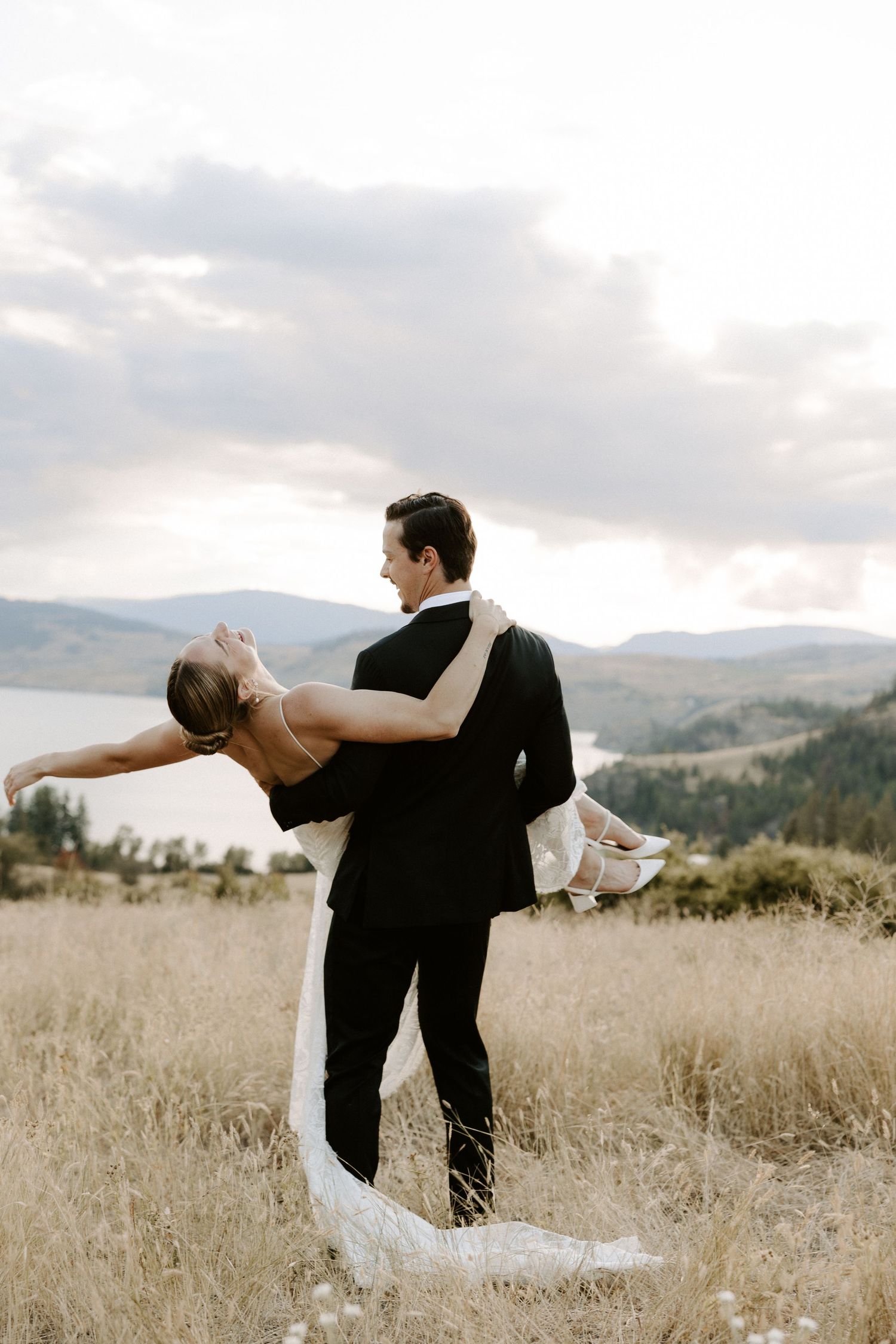 A couple in formal wedding attire dances together in golden field overlooking mountains and lake on cloudy day.