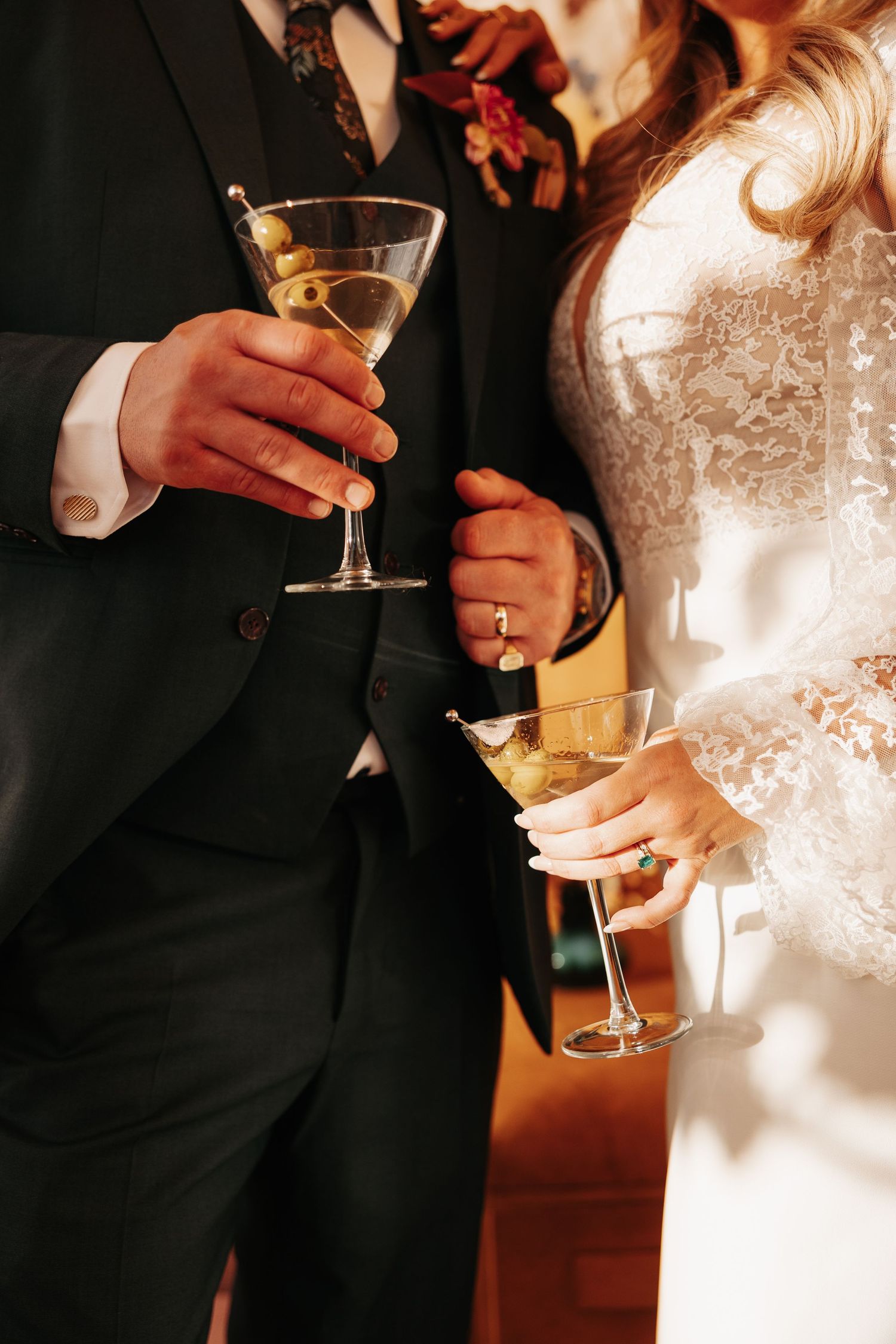 Close-up view of hands holding champagne glasses at a wedding reception with black suits and white dress visible.