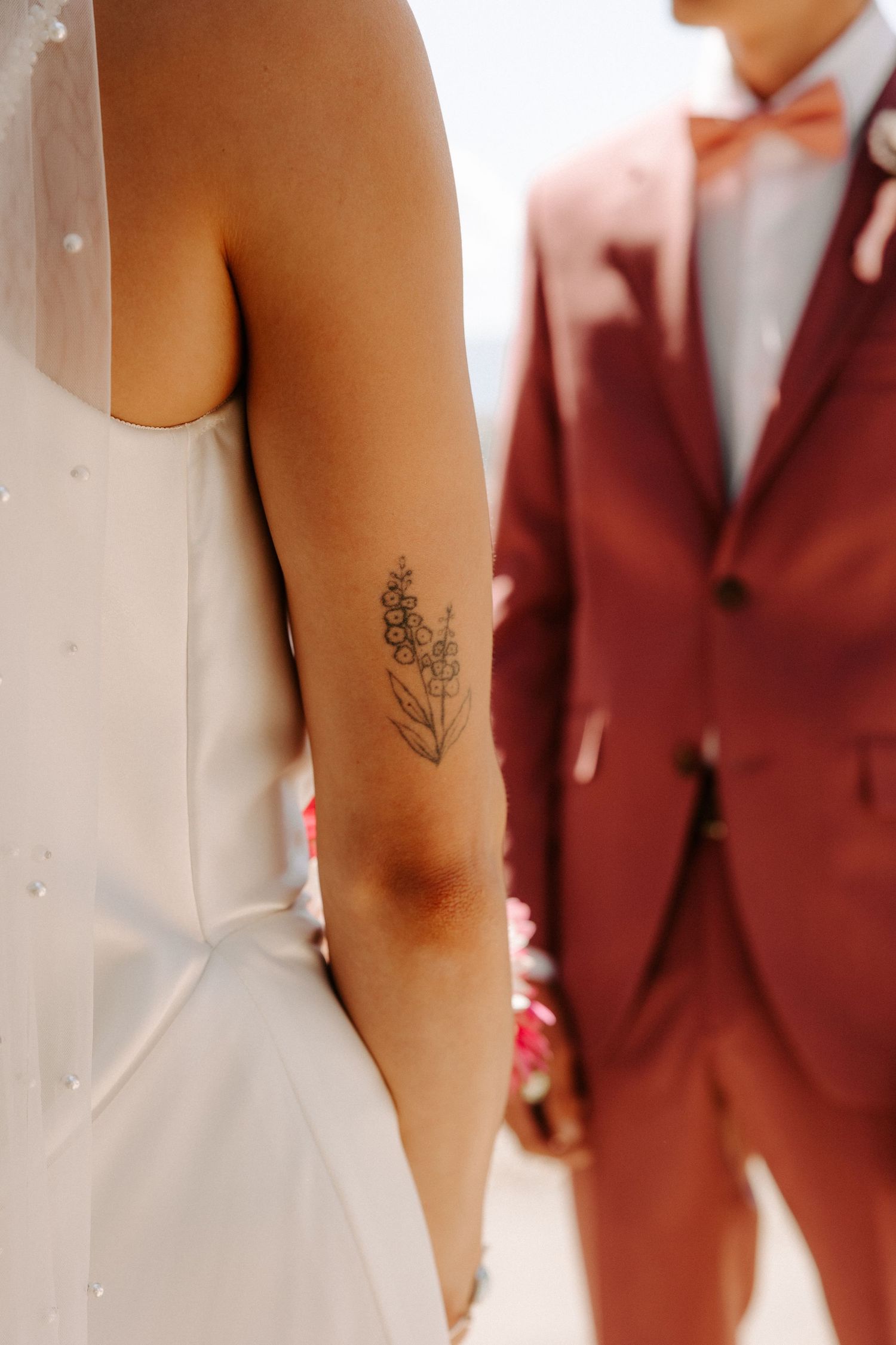 Wedding photo showing a delicate floral tattoo on a bride's arm next to someone wearing a burgundy suit.
