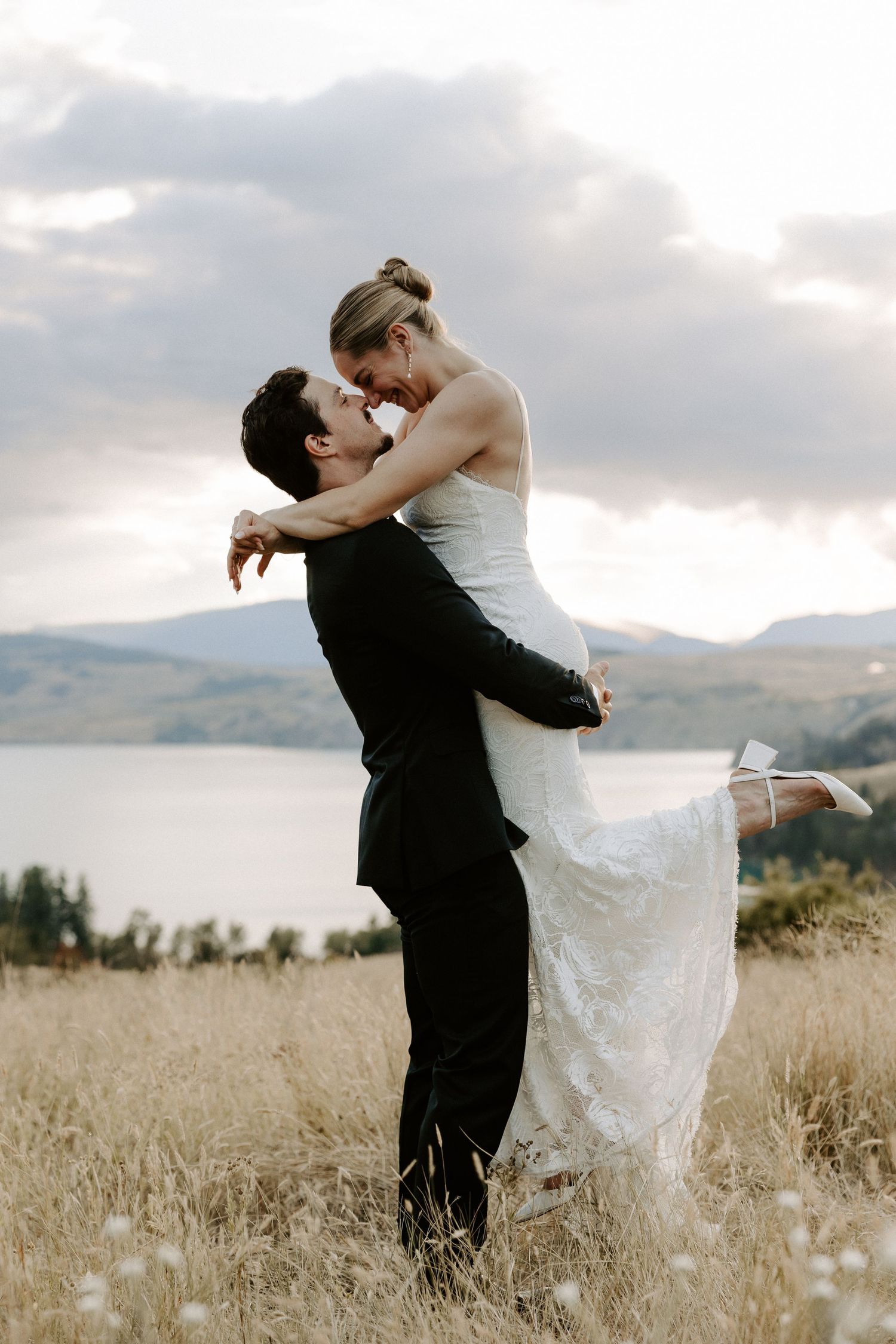 Romantic wedding couple embraces in tall grass field with mountains in background during golden hour sunset.