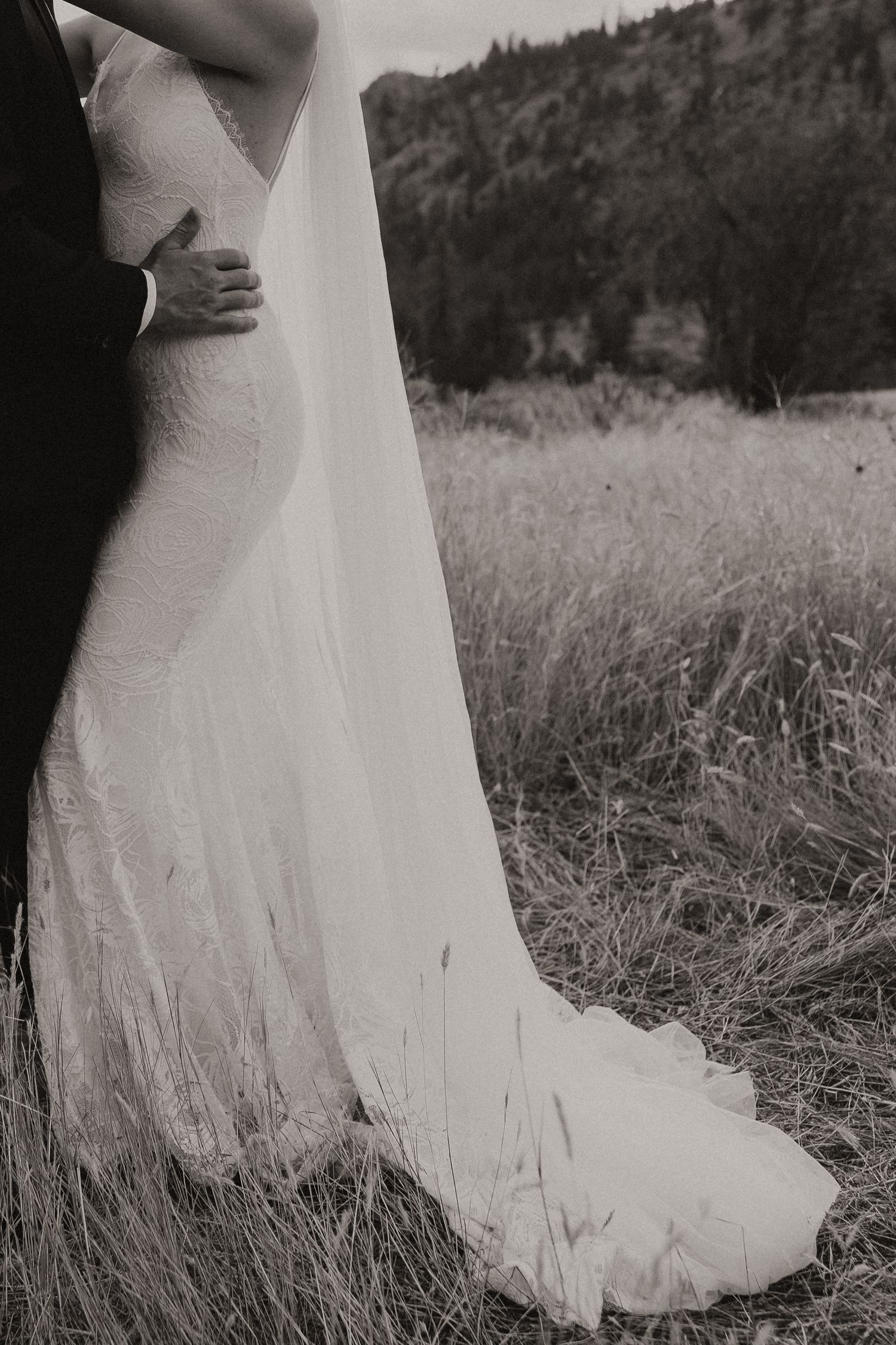 Black and white wedding dress detail photographed against tall grass with mountains in background at outdoor venue.
