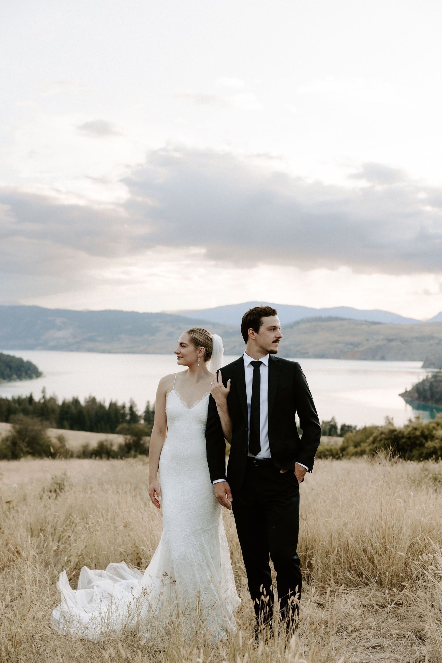 A newlywed couple stands in a scenic mountain meadow overlooking a lake during their wedding photoshoot.