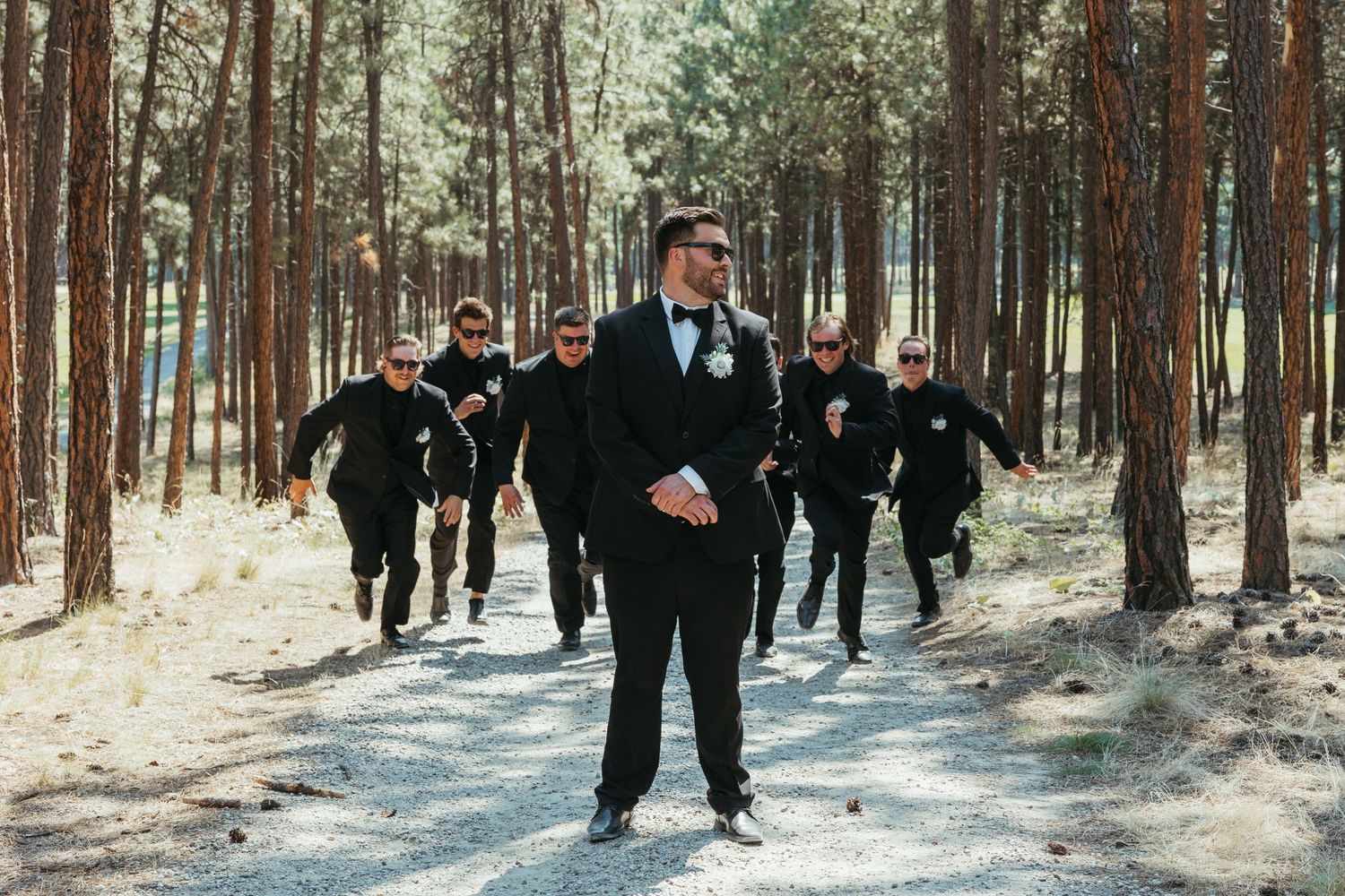 A wedding party group walks through a snowy pine forest with the groom leading in front.