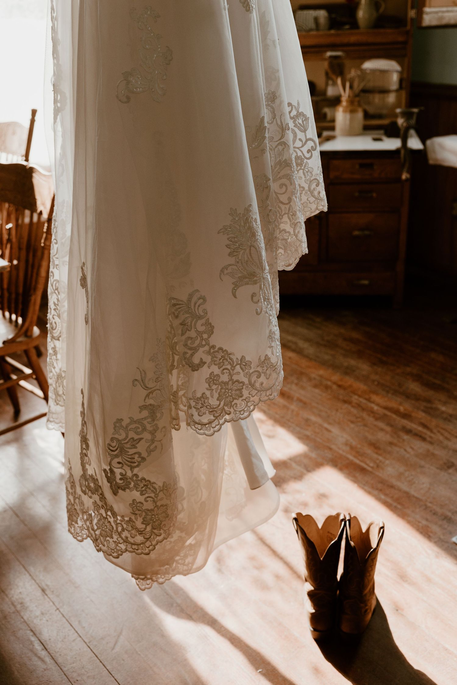 Wedding dress with lace detail hanging with cowboy boots in rustic room.