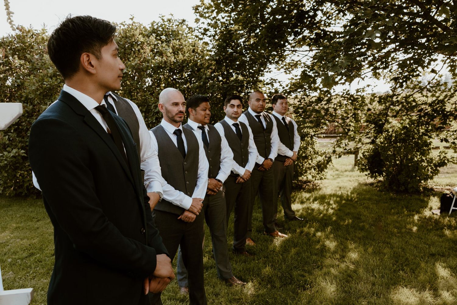 Groomsmen lined up outdoors wearing matching vests and formal attire.