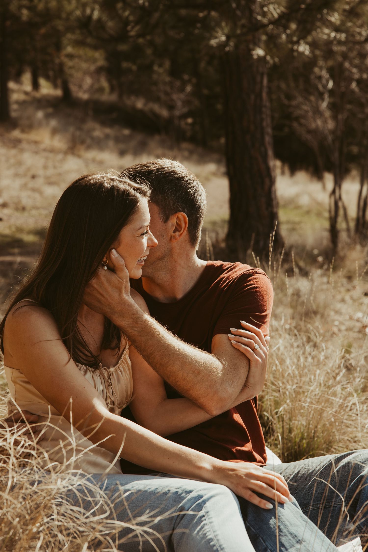 Intimate couple sharing tender moment while sitting in tall grass with sunlit forest backdrop.