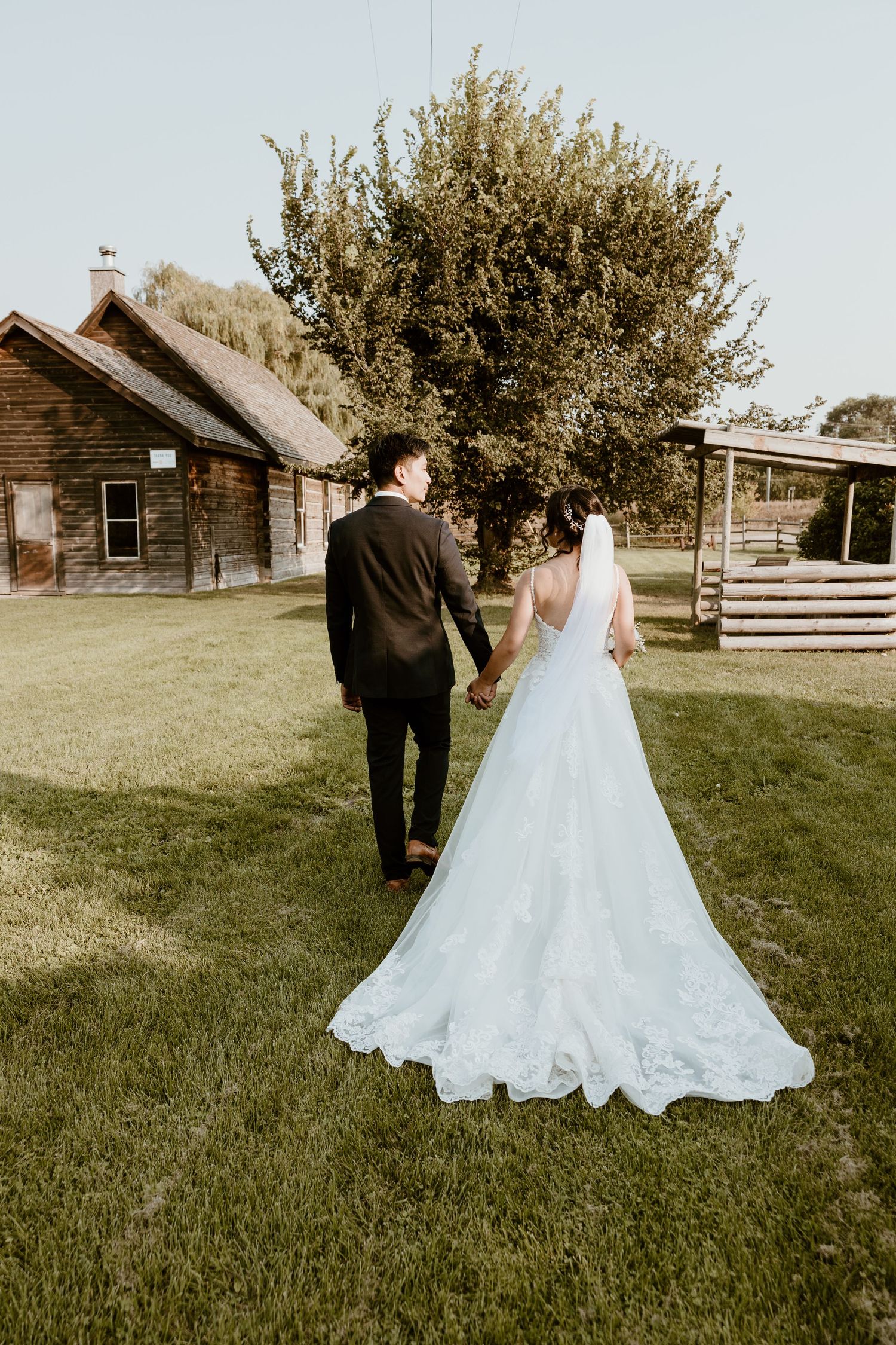 Bride and groom holding hands walking near rustic wooden buildings.
