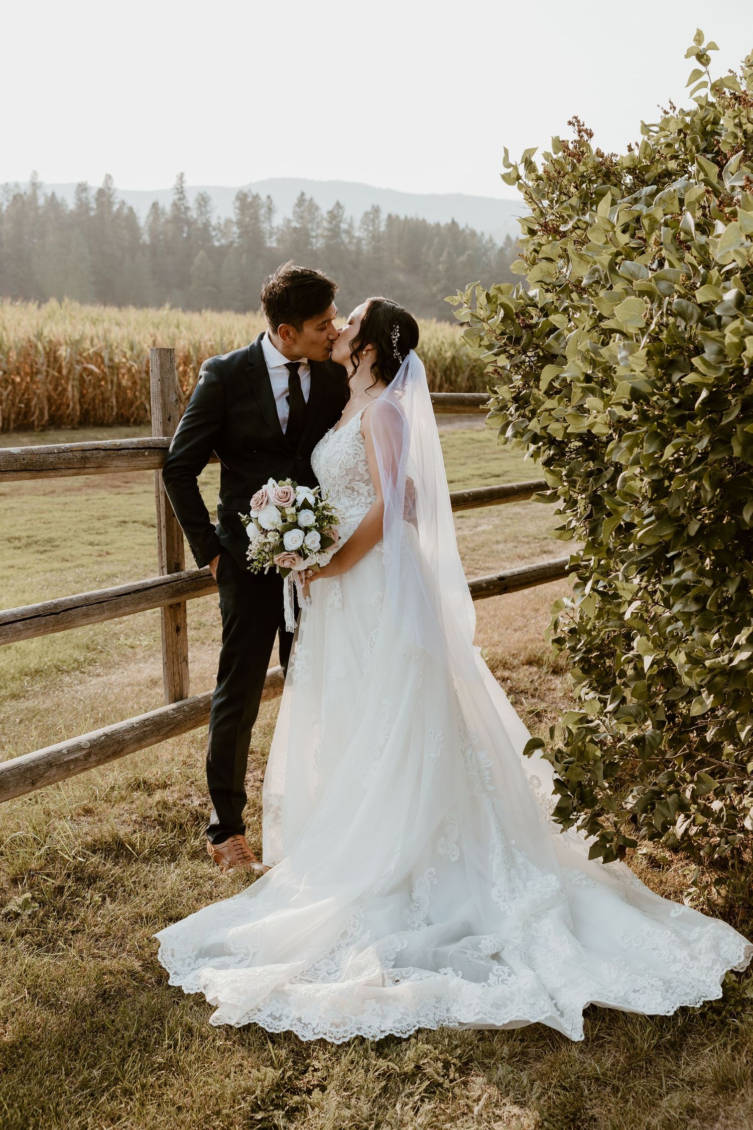 Newlyweds share a romantic moment by a rustic fence during golden hour with mountains in the background.