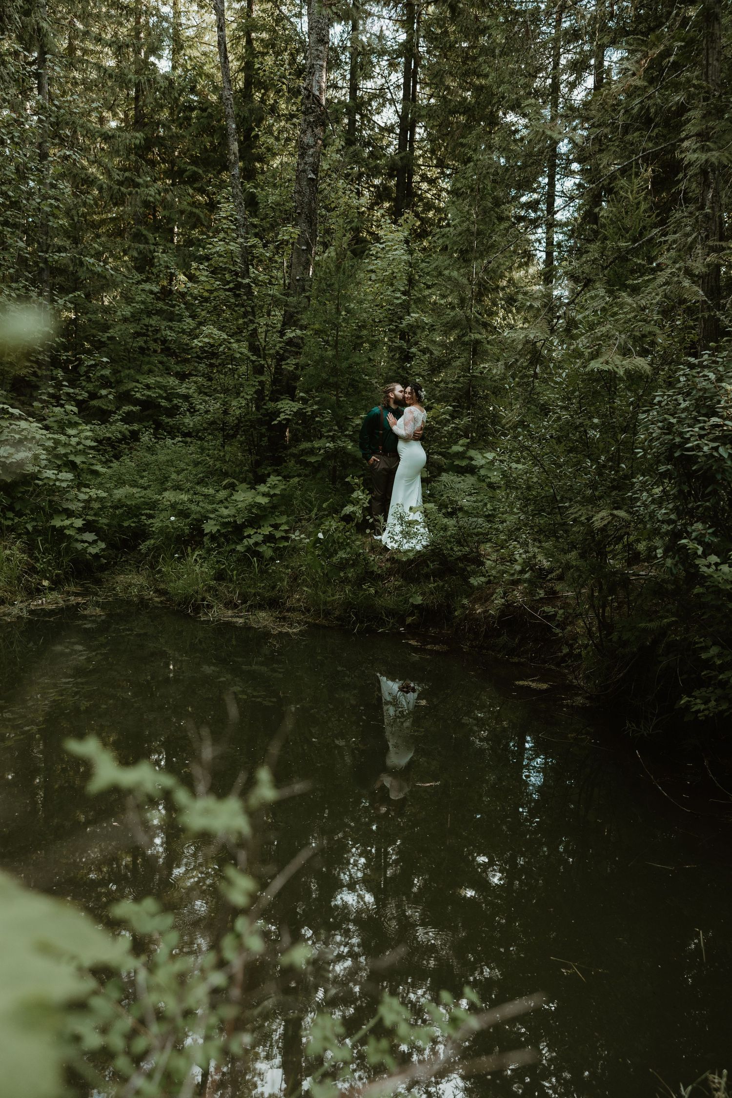 A wedding couple shares an intimate moment by a reflective forest pond.