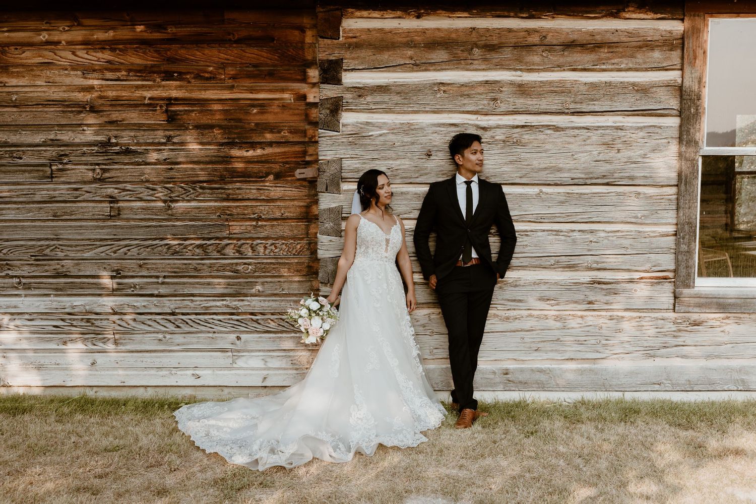 Wedding couple poses against weathered wooden barn wall in elegant attire.