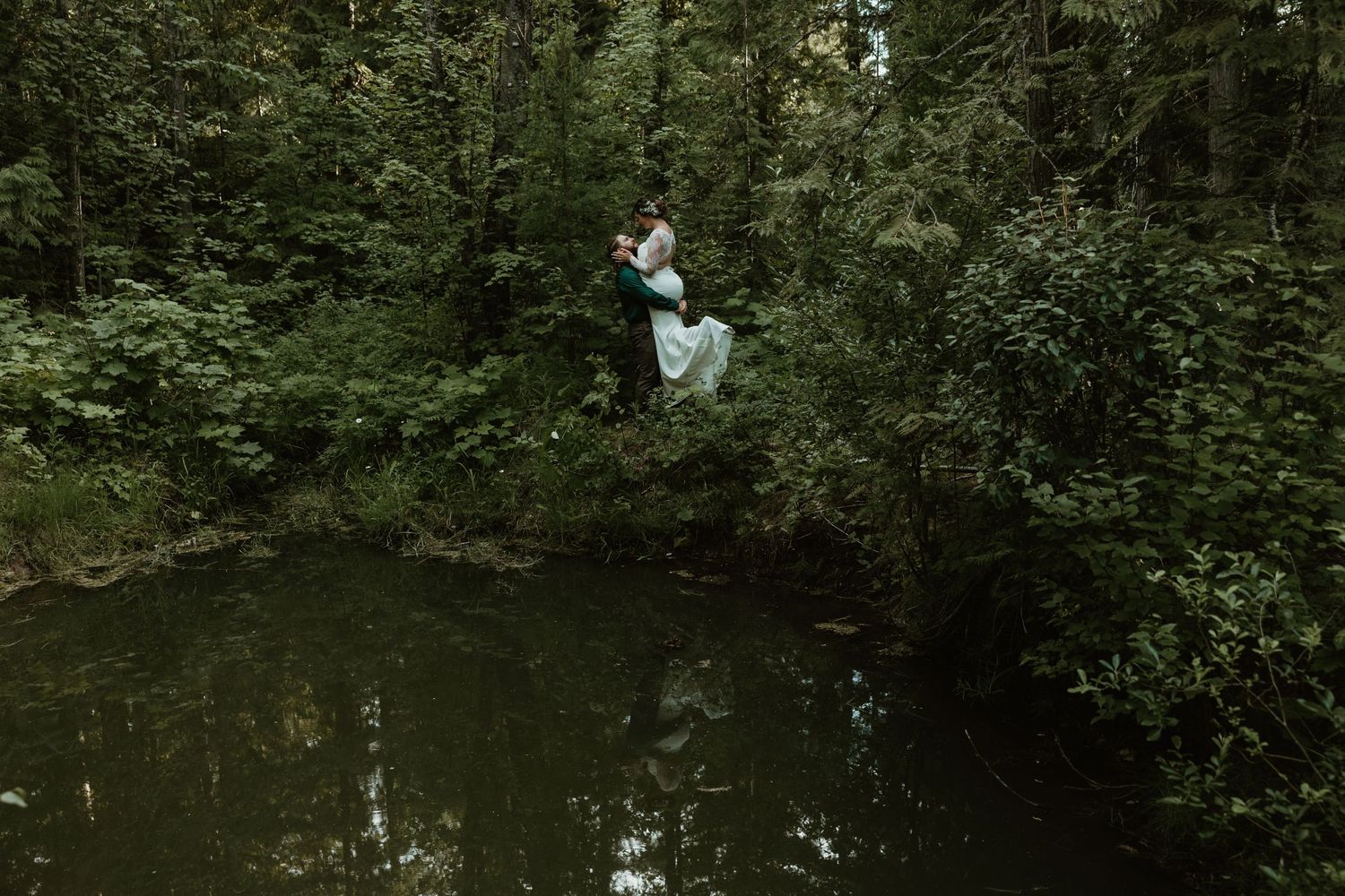 A couple in wedding attire sits playfully on a log above a forest pond.