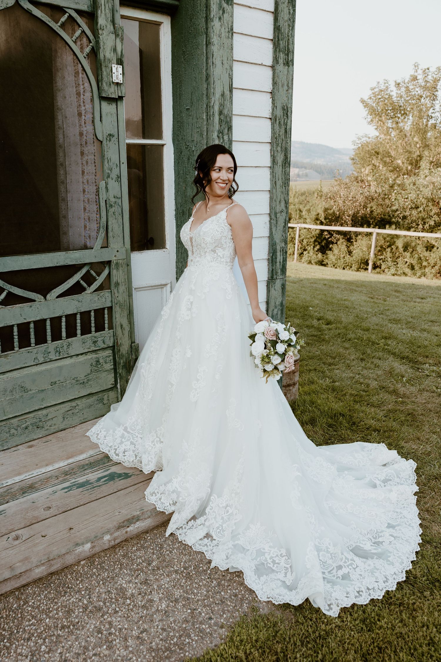 Bride in lace wedding gown with long train poses on vintage porch.