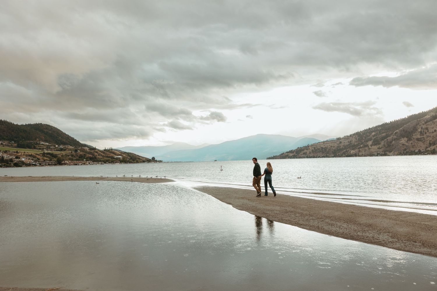 Two silhouettes stand on a misty beach with mountains in the background.