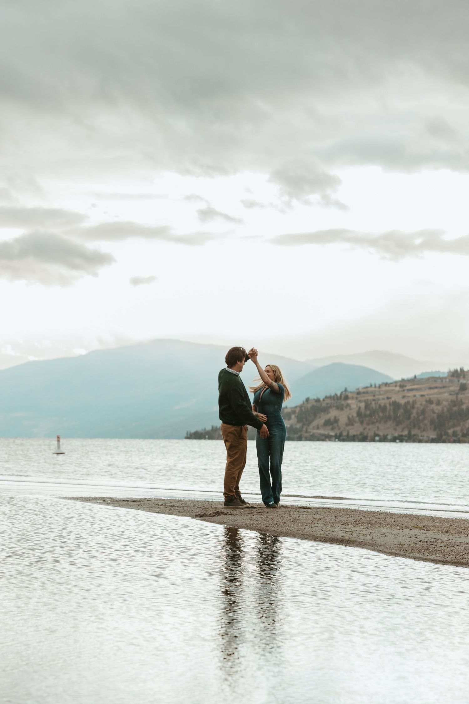 A couple stands together on a beach shore with mountains and cloudy skies in the background.
