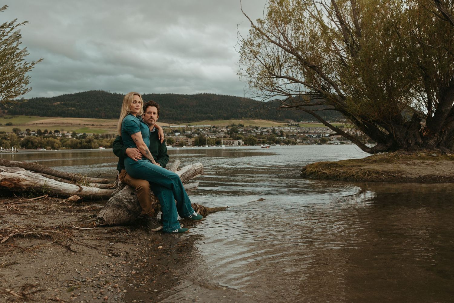 A couple sits on driftwood by a serene river with mountains in the distance.