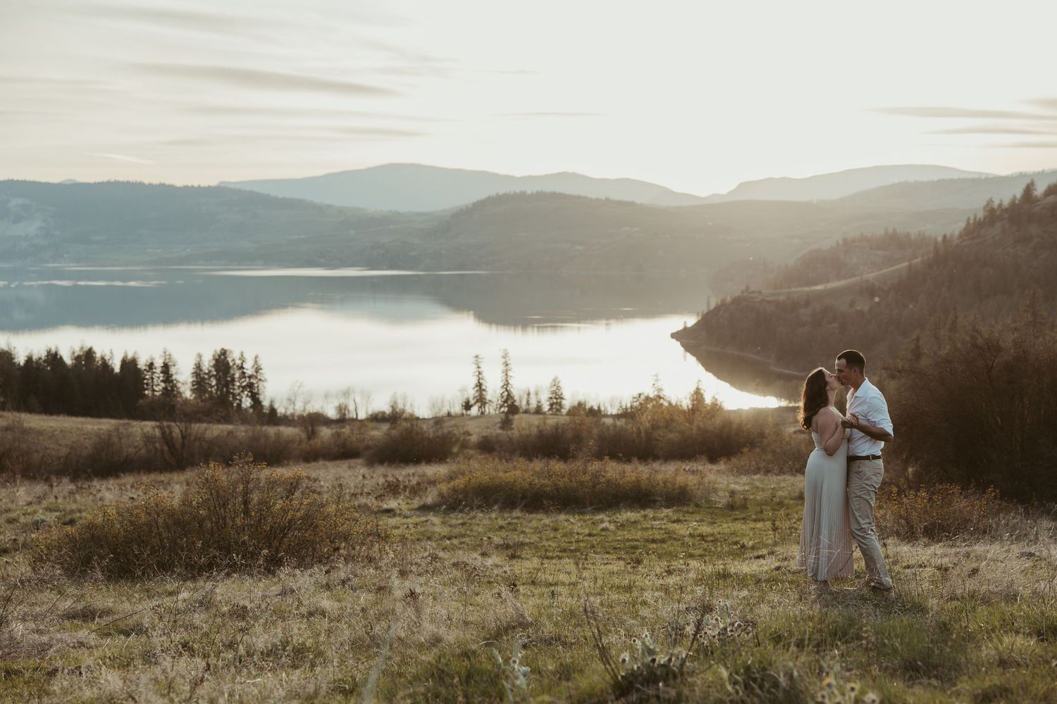 A couple shares an intimate moment on a hilltop overlooking a misty lake at sunset.