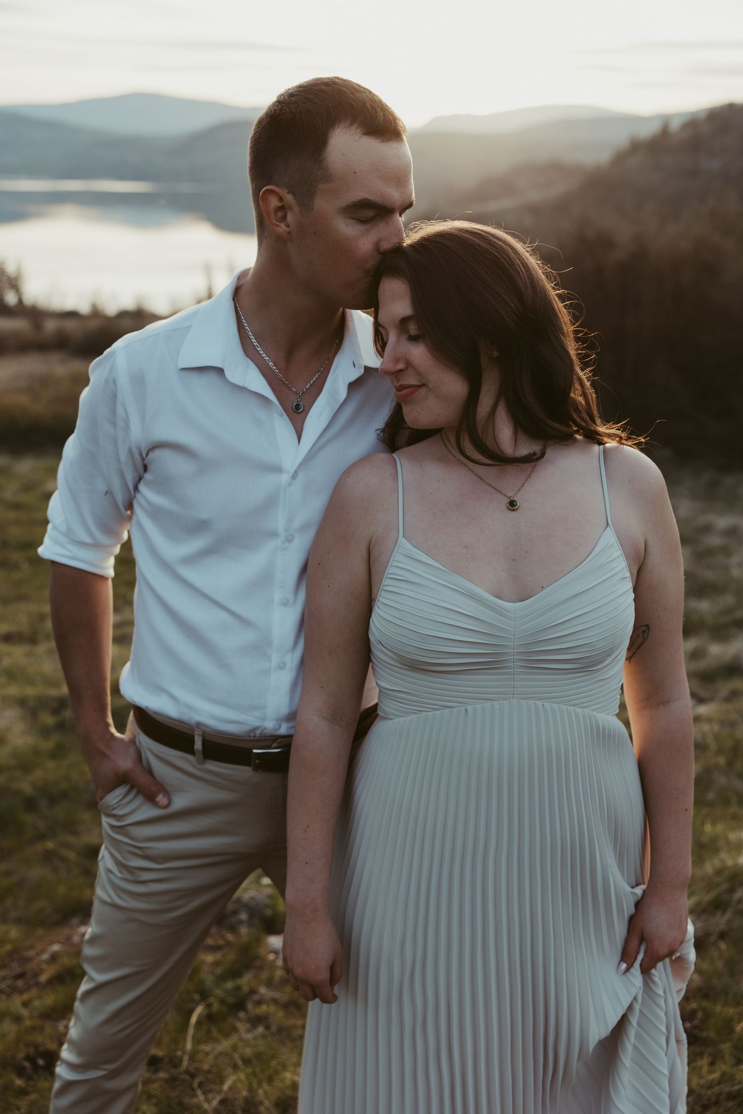 A romantic sunset engagement photo of a couple posing together on a scenic hilltop overlooking a lake.