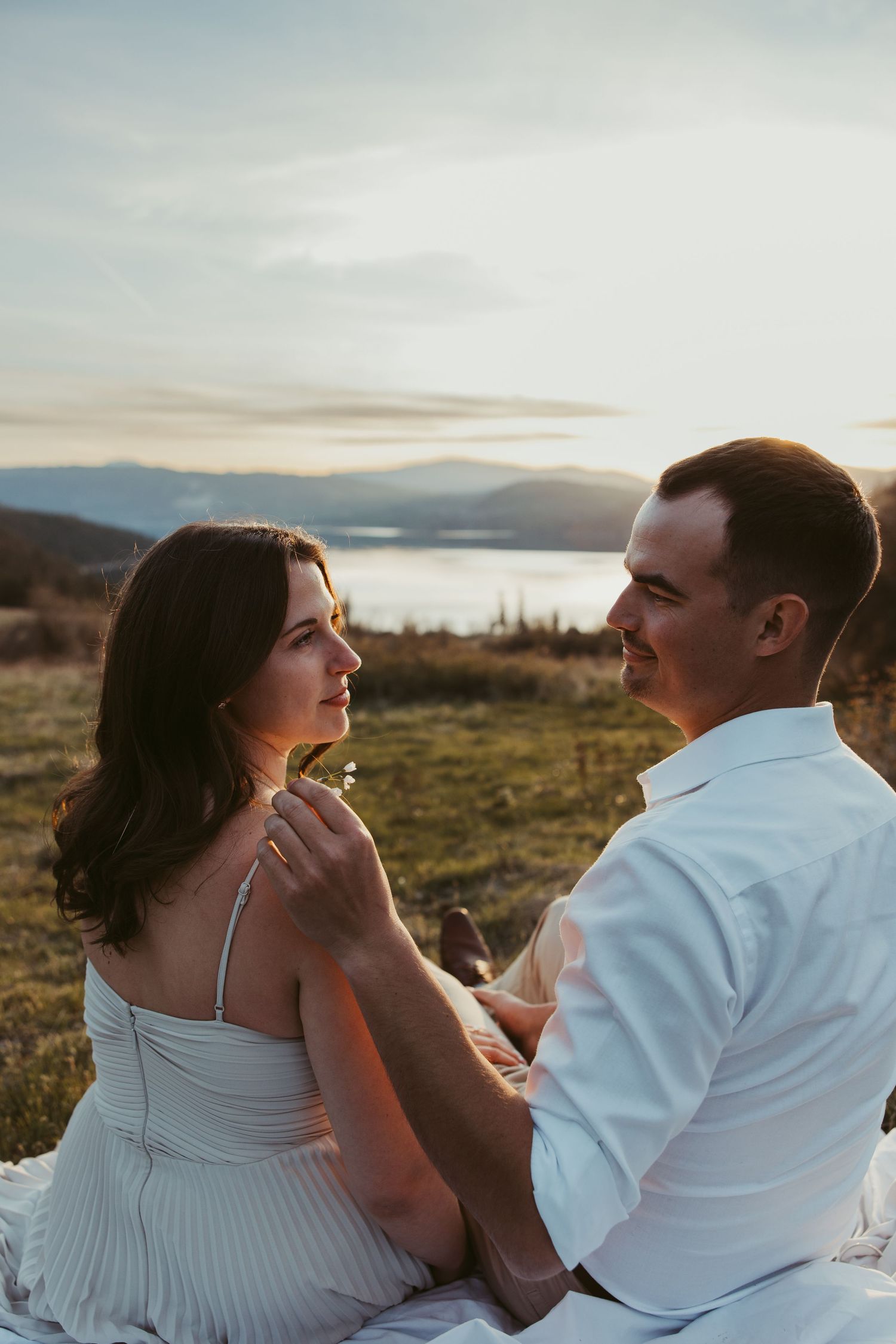 A romantic outdoor photoshoot featuring a couple embracing on a hillside with a lake view at sunset.