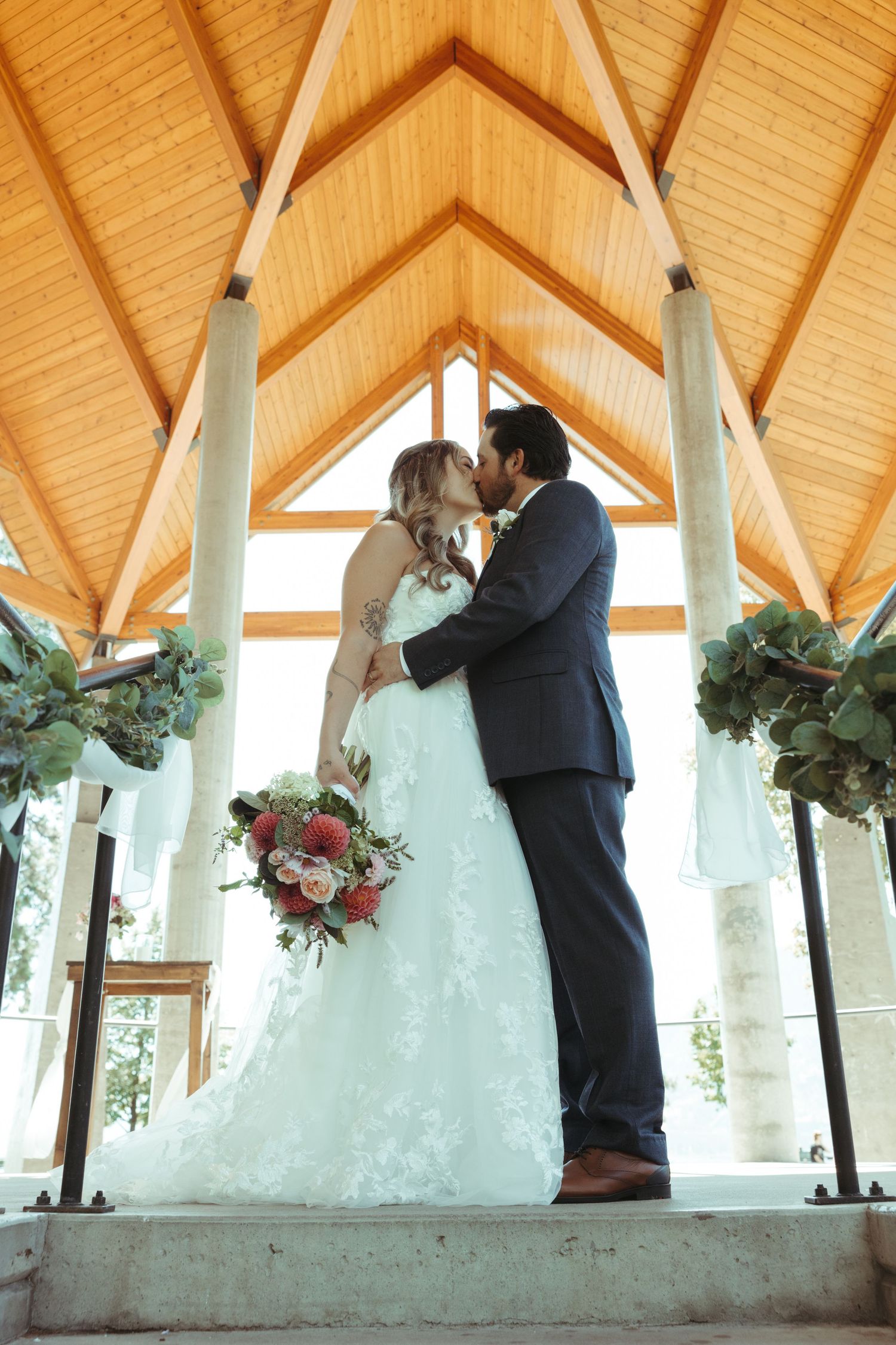 Newlyweds share a kiss under a wooden pavilion decorated with floral arrangements.