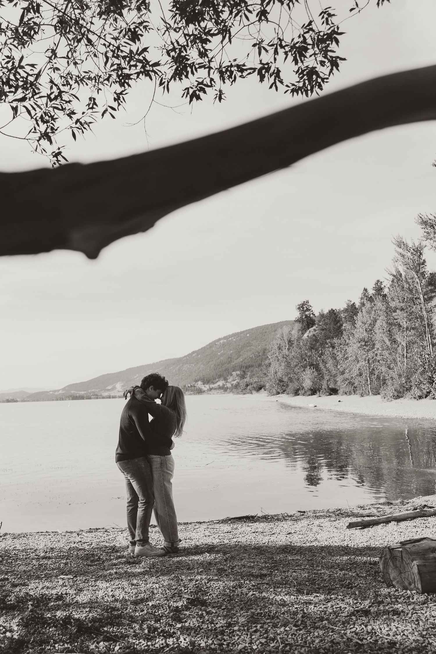 A romantic couple embraces by a serene lakeside beach with mountains in the background.