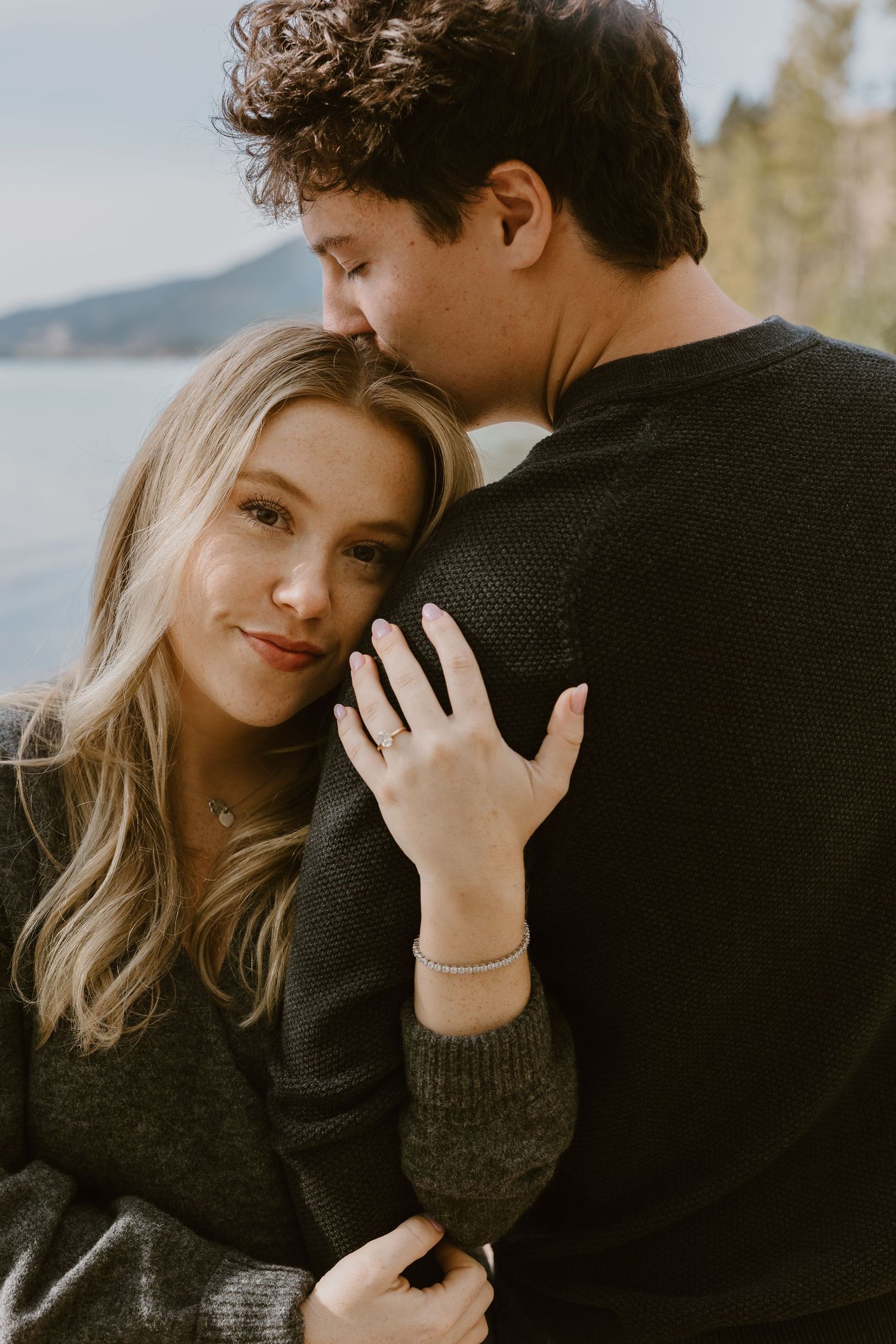 An intimate embrace between a couple wearing dark sweaters against a waterfront backdrop.
