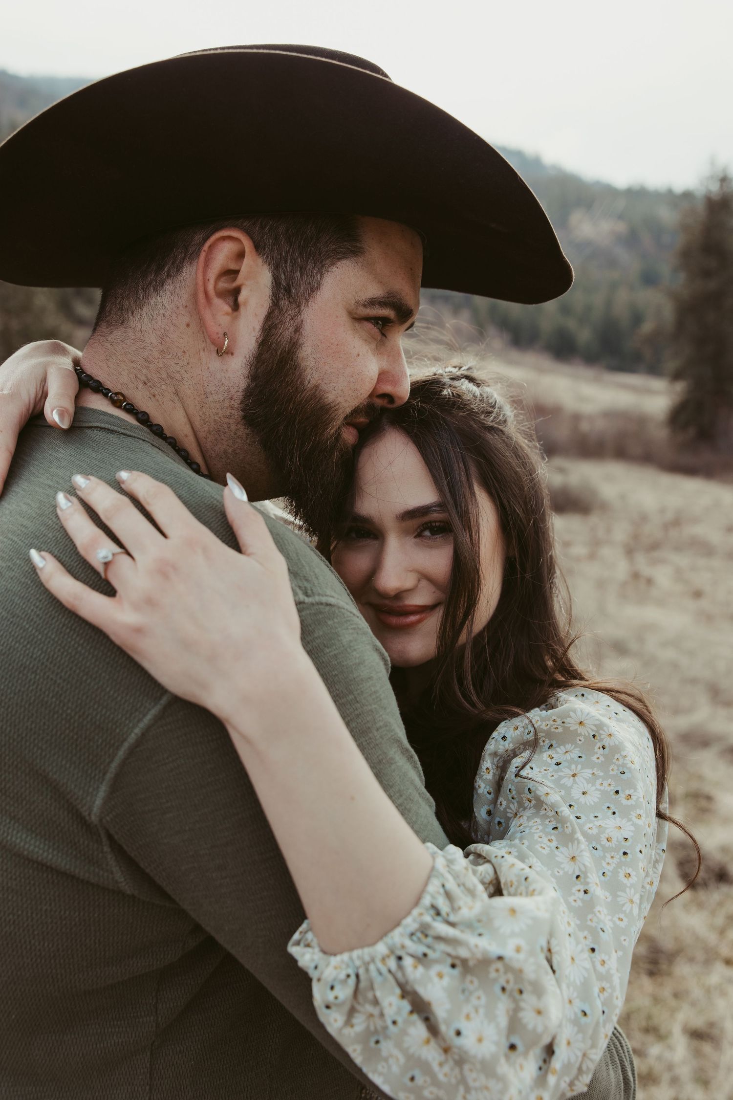 A romantic couple embraces in a field wearing western attire with a cowboy hat and floral dress.