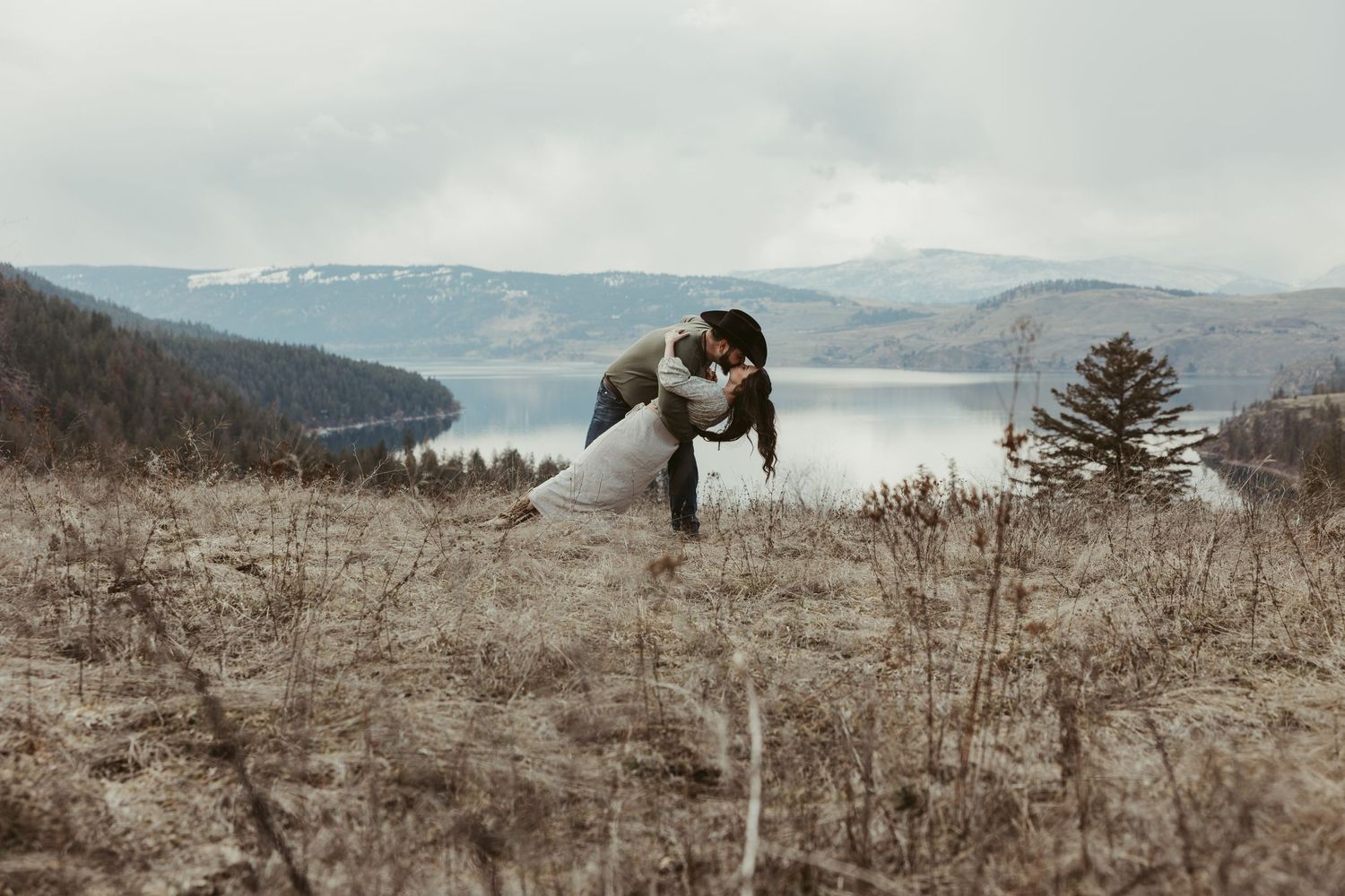 A playful couple poses dramatically on a hilltop with a lake view in the background.