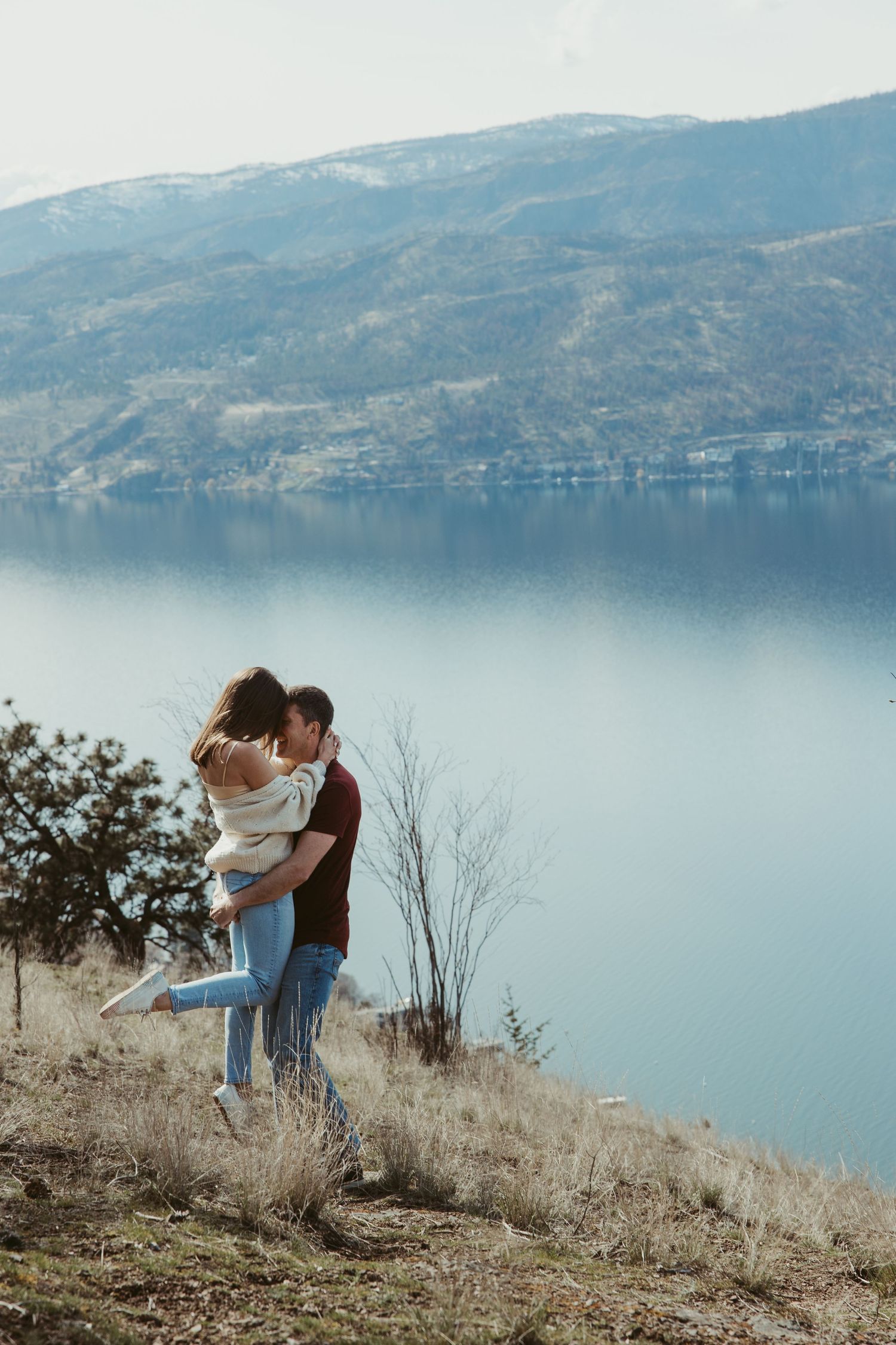 Couple sharing joyful embrace on hillside overlooking serene lake and mountains.