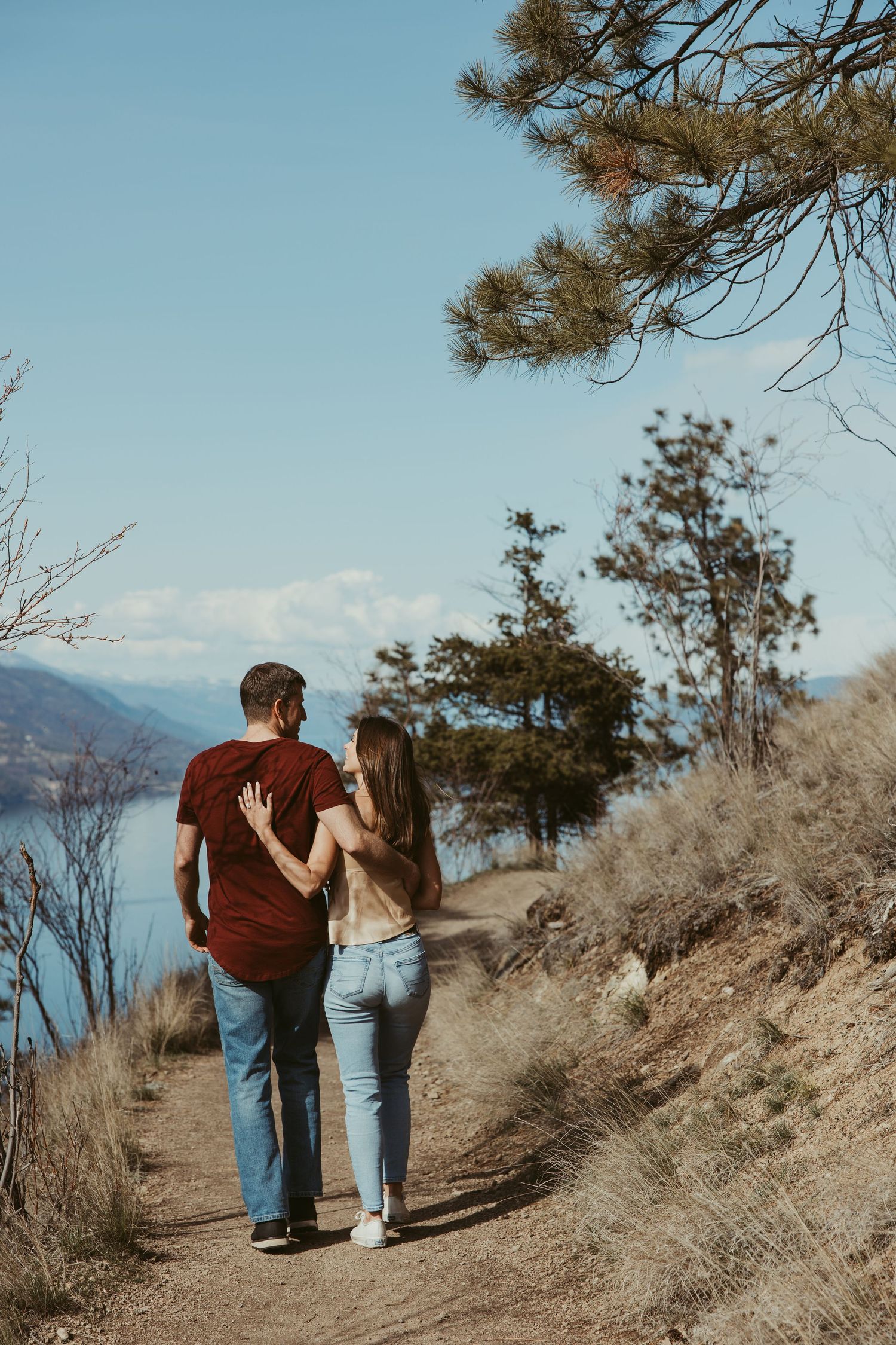 Couple walking together on dirt trail with lake and mountain views in background.