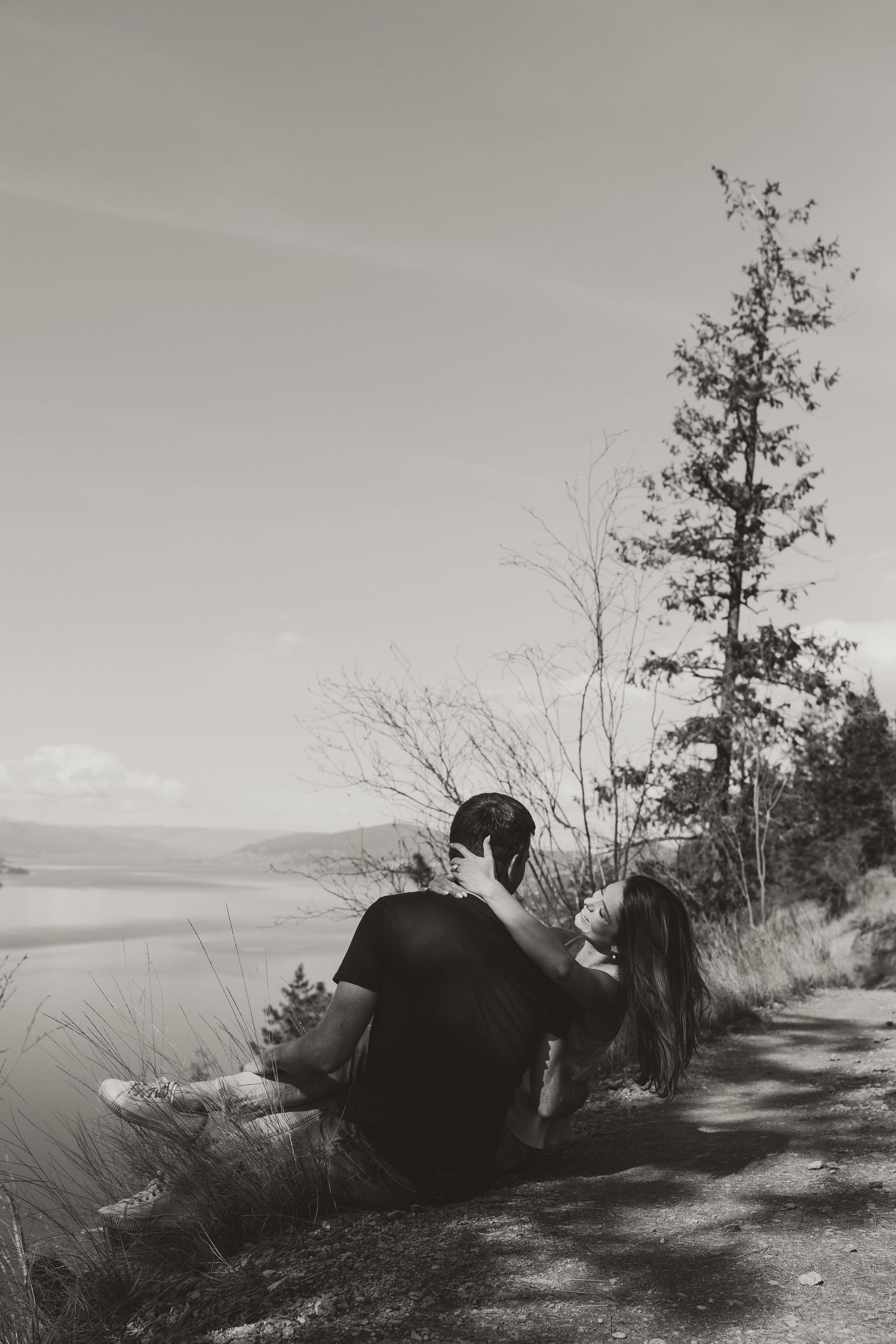 A couple sits together enjoying a scenic lake view from a hillside path in black and white.