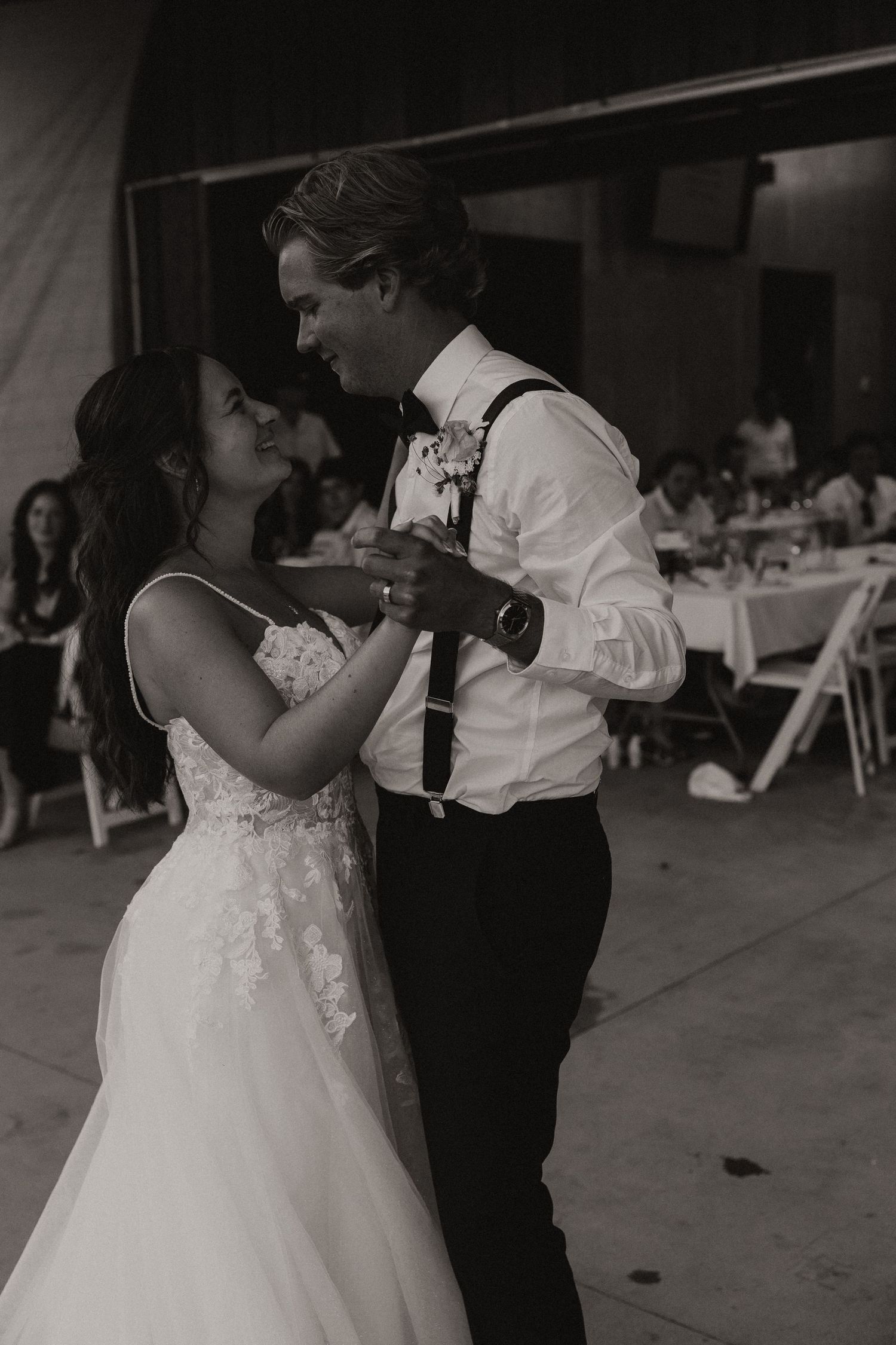 A bride and groom share an intimate moment during their first dance at a wedding reception venue with tables in background.