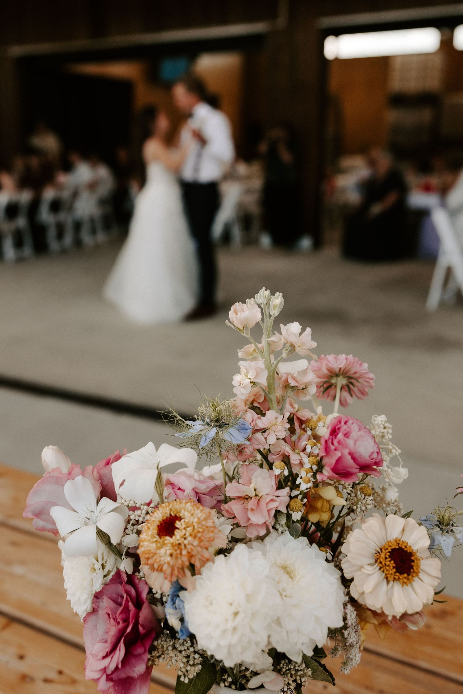 Beautiful floral centerpiece with pink and white blooms on wooden table as couple dances in blurred background at wedding reception.