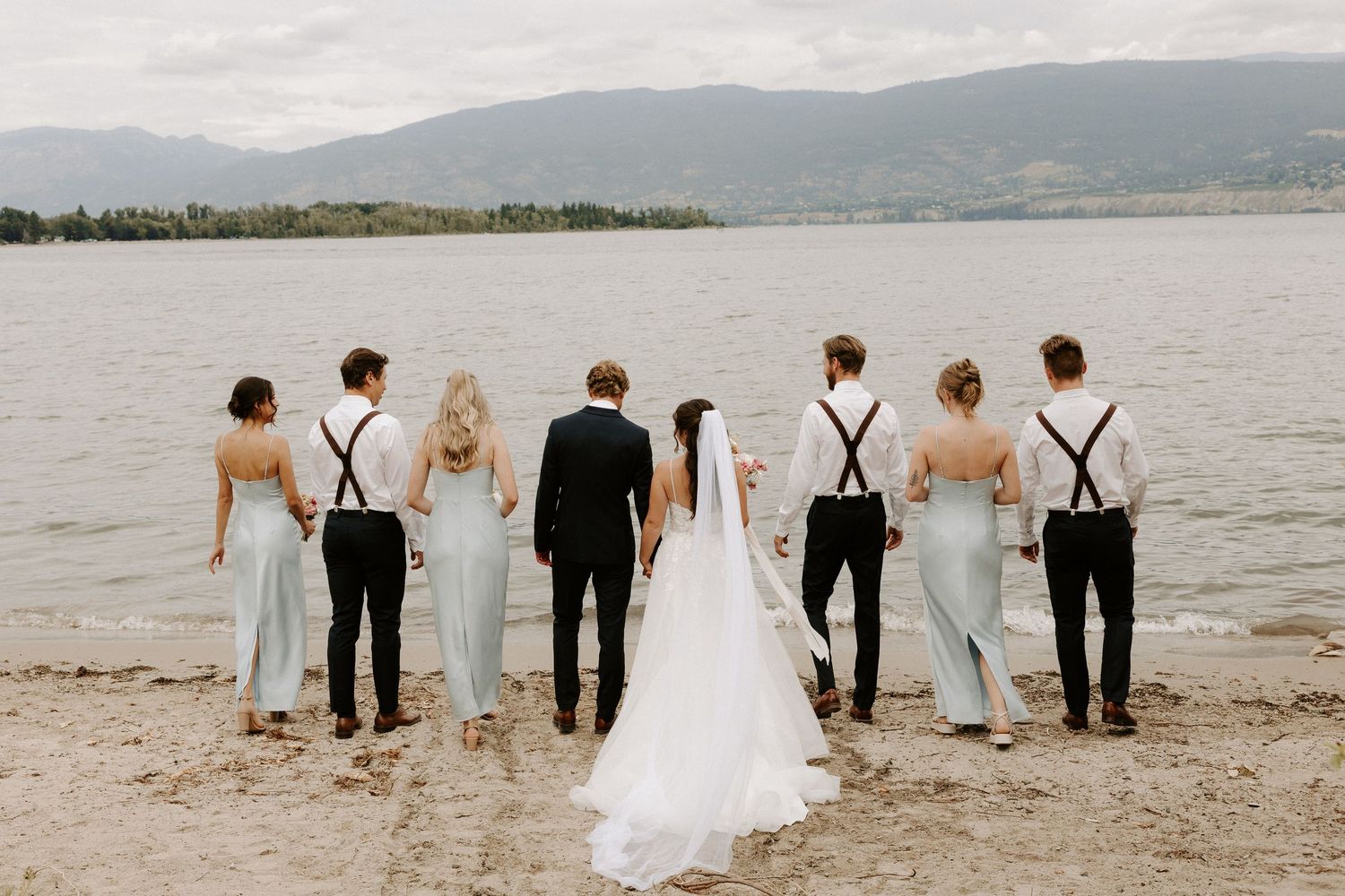 Wedding party walks along shoreline with mountain backdrop as they pose for scenic beach photo.