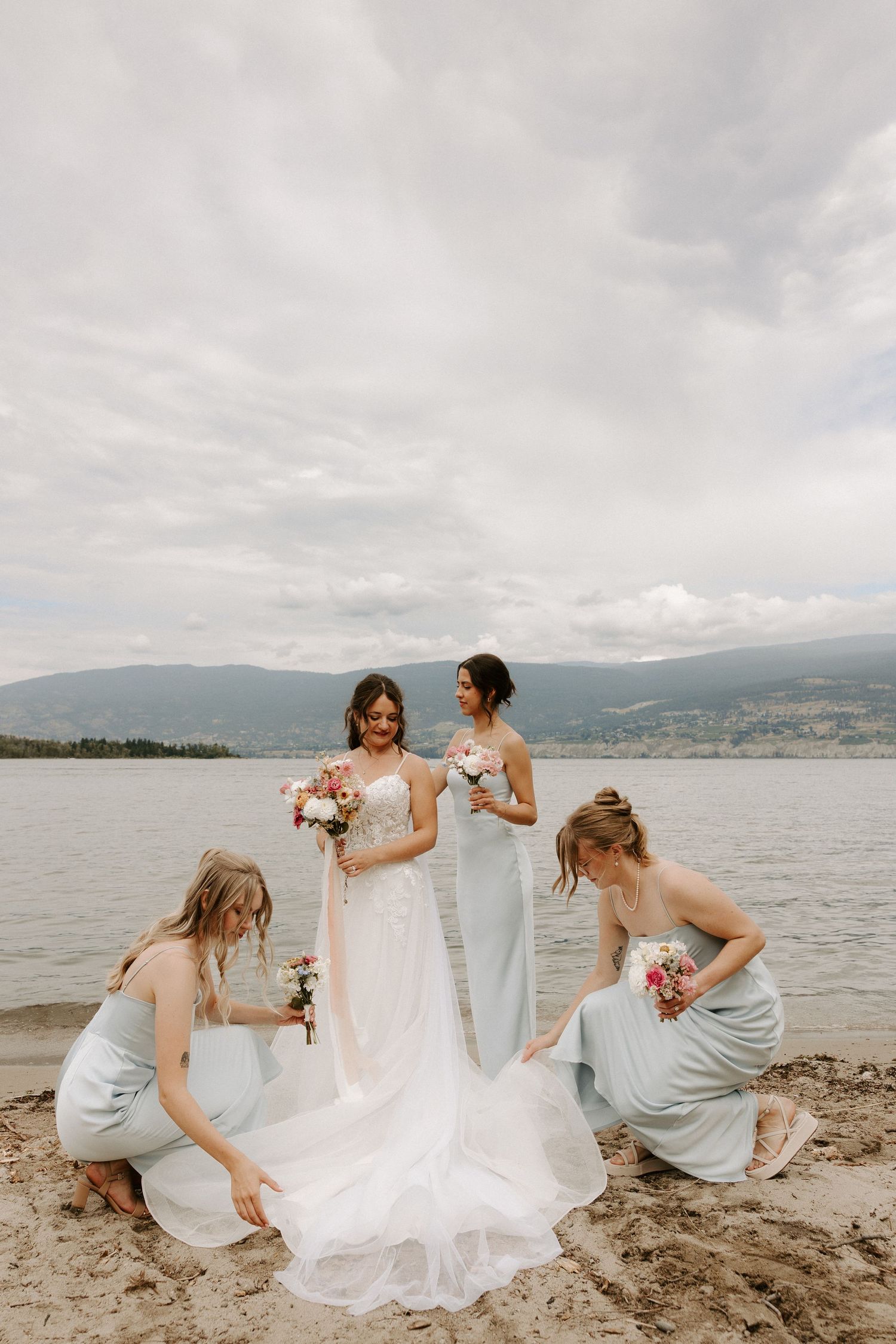 Wedding party in light blue dresses helps with bride's gown on lakeside beach with mountains in background.